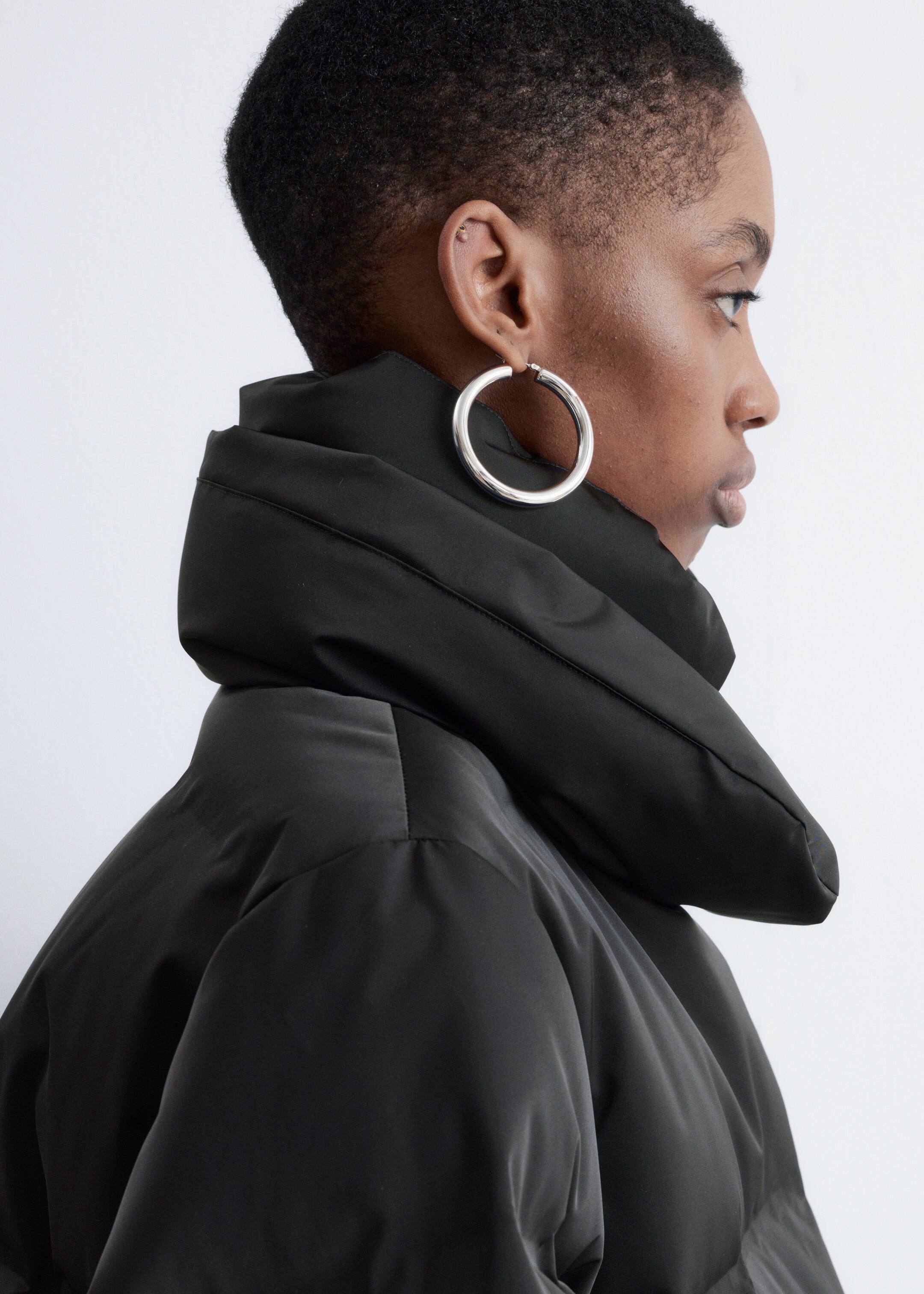 View larger image: A black puffer jacket is being presented from a side angle, featuring a high, padded stand-up collar. The jacket's shoulder area is appearing rounded and voluminous.