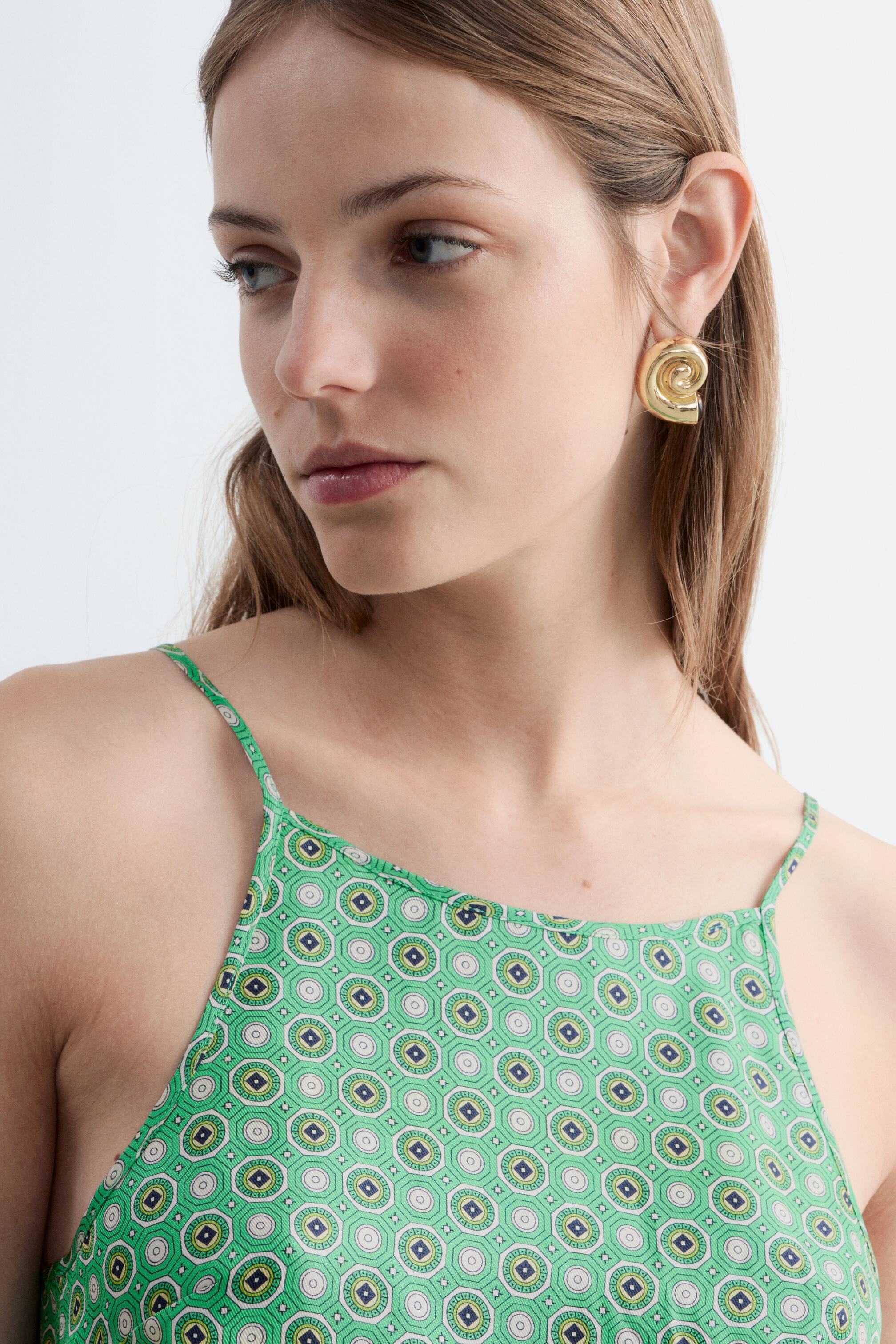Ver imagem maior: Um top verde estampado apresenta uma gola alta e finas alças de espaguete, exibindo um design geométrico com motivos octogonais e circulares repetidos em tons de verde, branco e azul escuro.