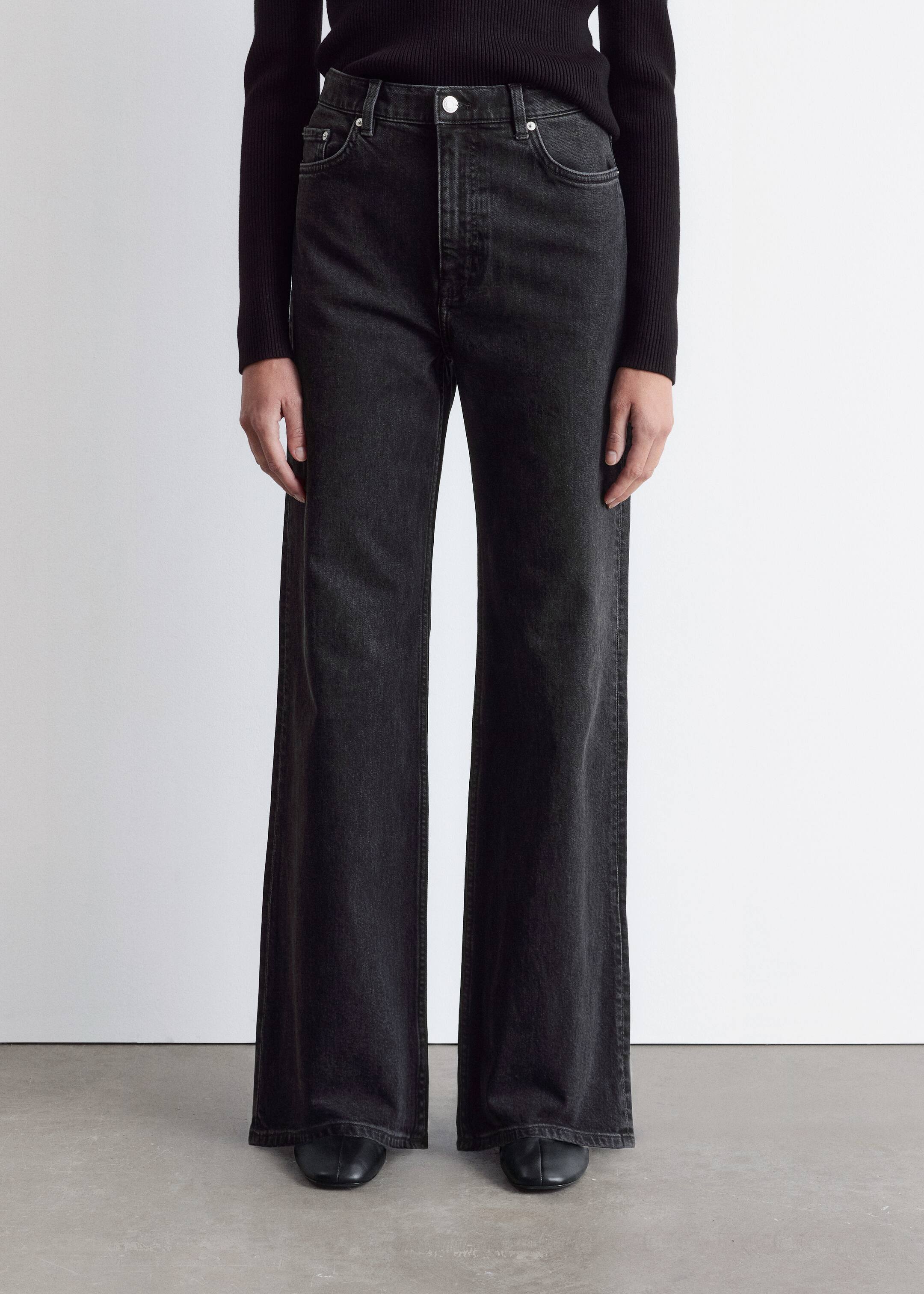 View larger image: Washed black high-waisted, wide-leg jeans are being presented from a front angle. The jeans are featuring a single button closure, a zipper fly, and two front pockets with visible metal rivets. Their wide legs are extending straight down, slightly pooling over black shoes, while a black ribbed top is being tucked into the waistband.