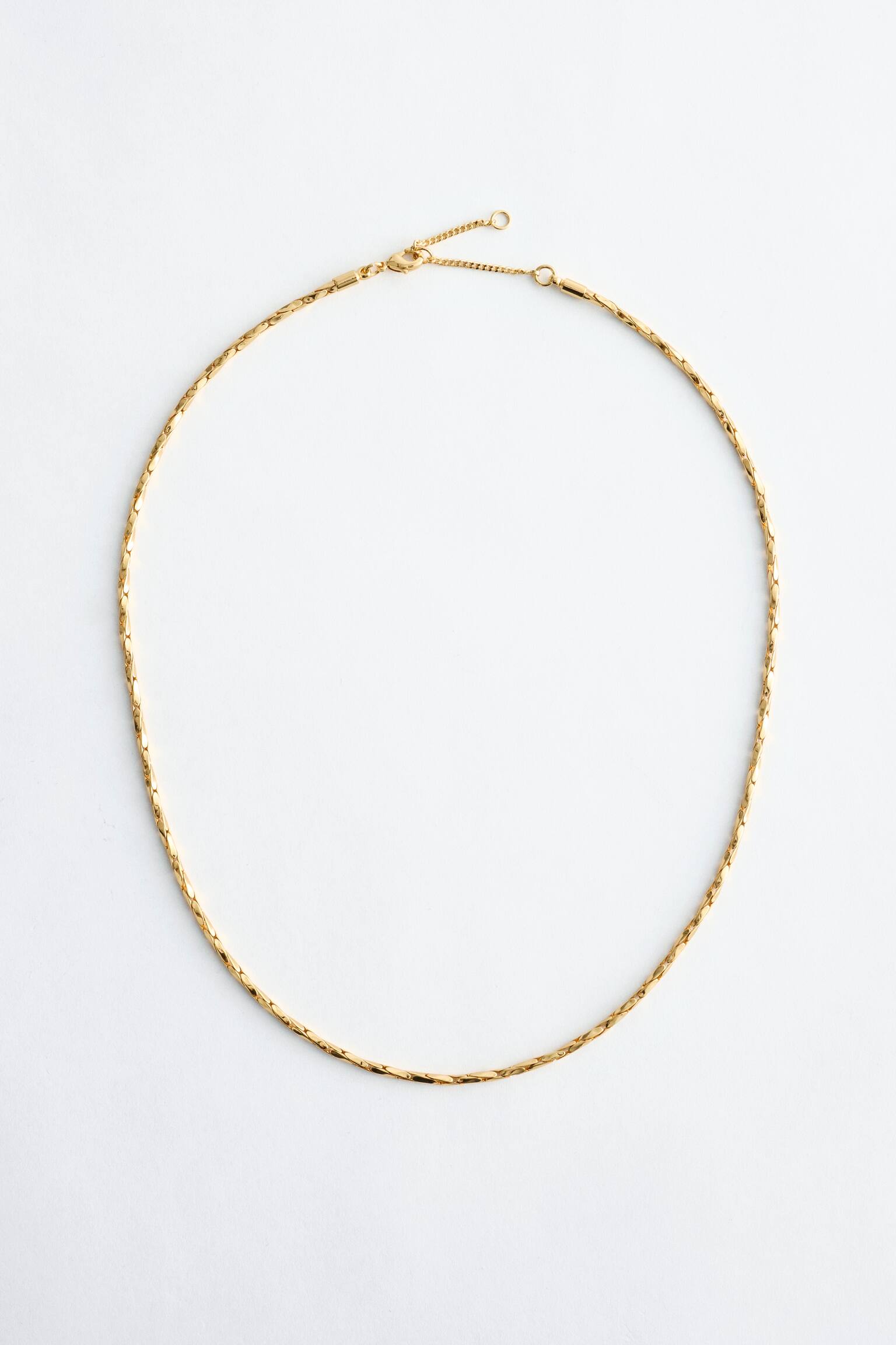 Een gepolijste, goudkleurige ketting toont zijn volledige lengte en vormt een open cirkel. Het beschikt over een flexibele ketting van korte, platte, onderling verbonden schakels en heeft een karabijnsluiting met een verlengketting aan de bovenkant. - 1
