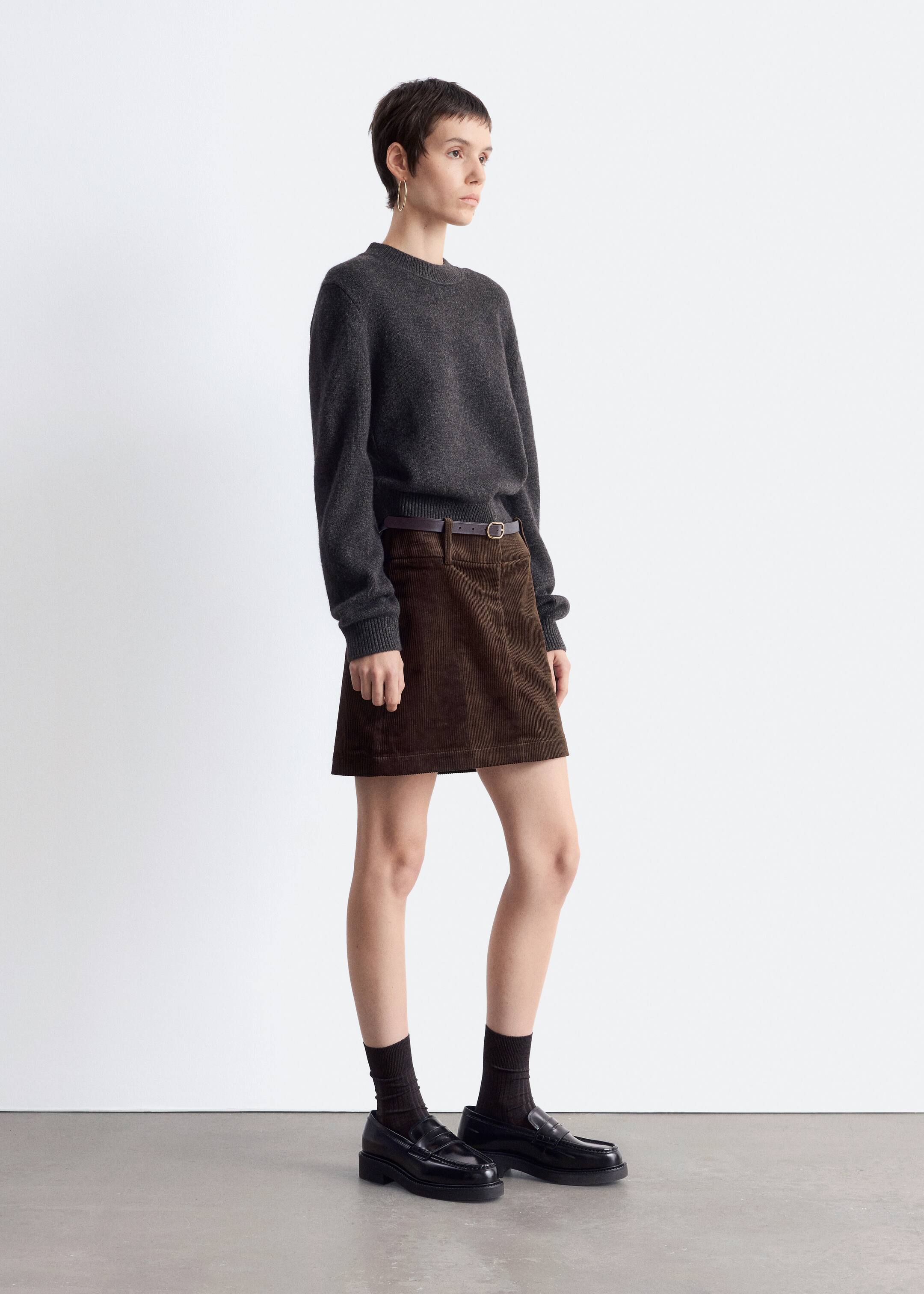 Größeres Bild ansehen: Ein dunkelgrauer kurzer Strickpullover mit Rundhalsausschnitt, gerippten Abschlüssen und sanft fallenden Schultern wird von dem Model in einer schmalen Passform getragen und mit einem braunen Cordrock kombiniert.
