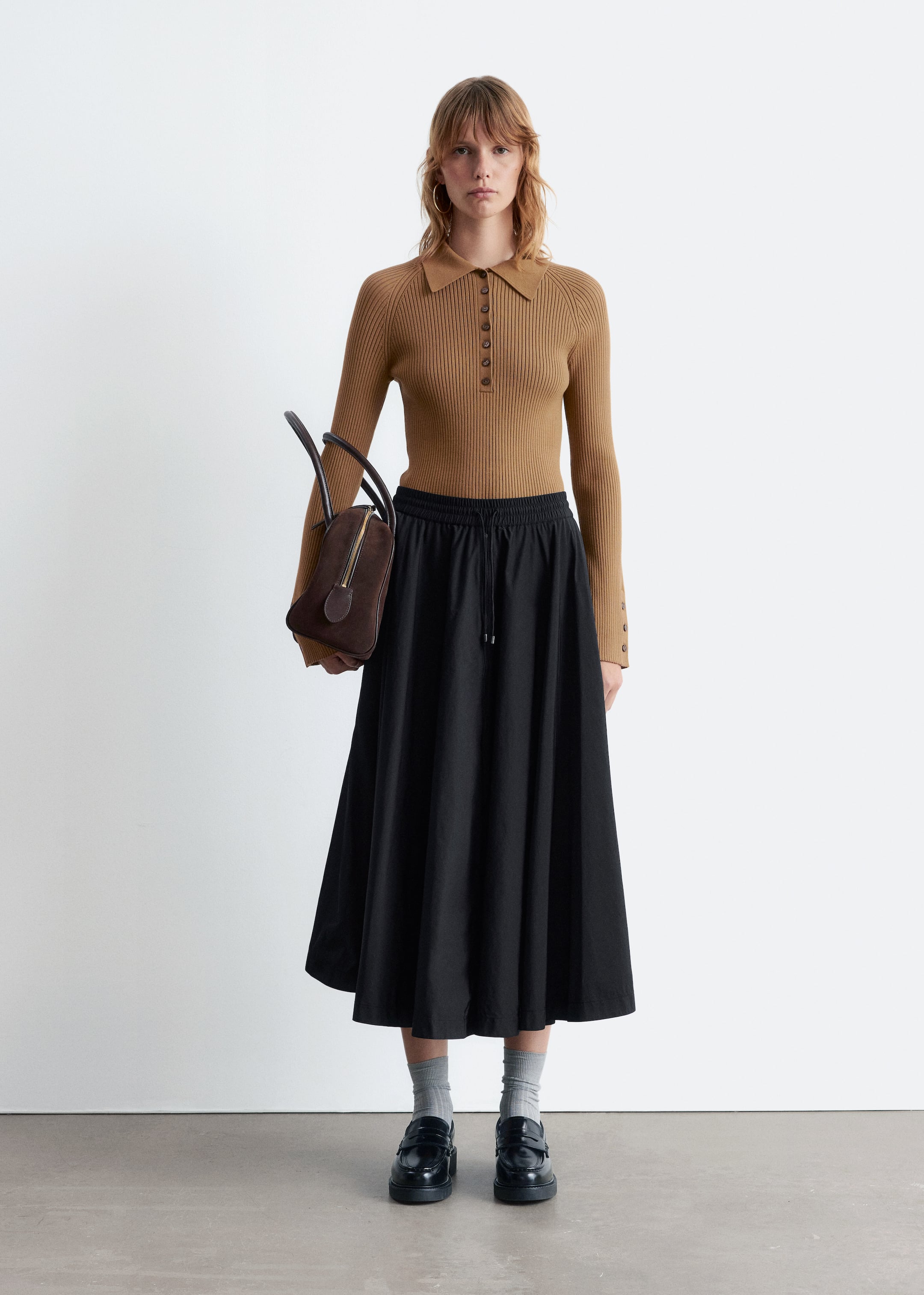 Agrandir l'image: Un haut polo en maille côtelée couleur camel avec un col pointu et une patte de boutonnage est porté, associé à une jupe noire de longueur midi avec une taille élastiquée à cordon de serrage. Des mocassins noirs épais sont portés avec des chaussettes côtelées gris clair, et un sac à main en daim marron foncé est tenu.