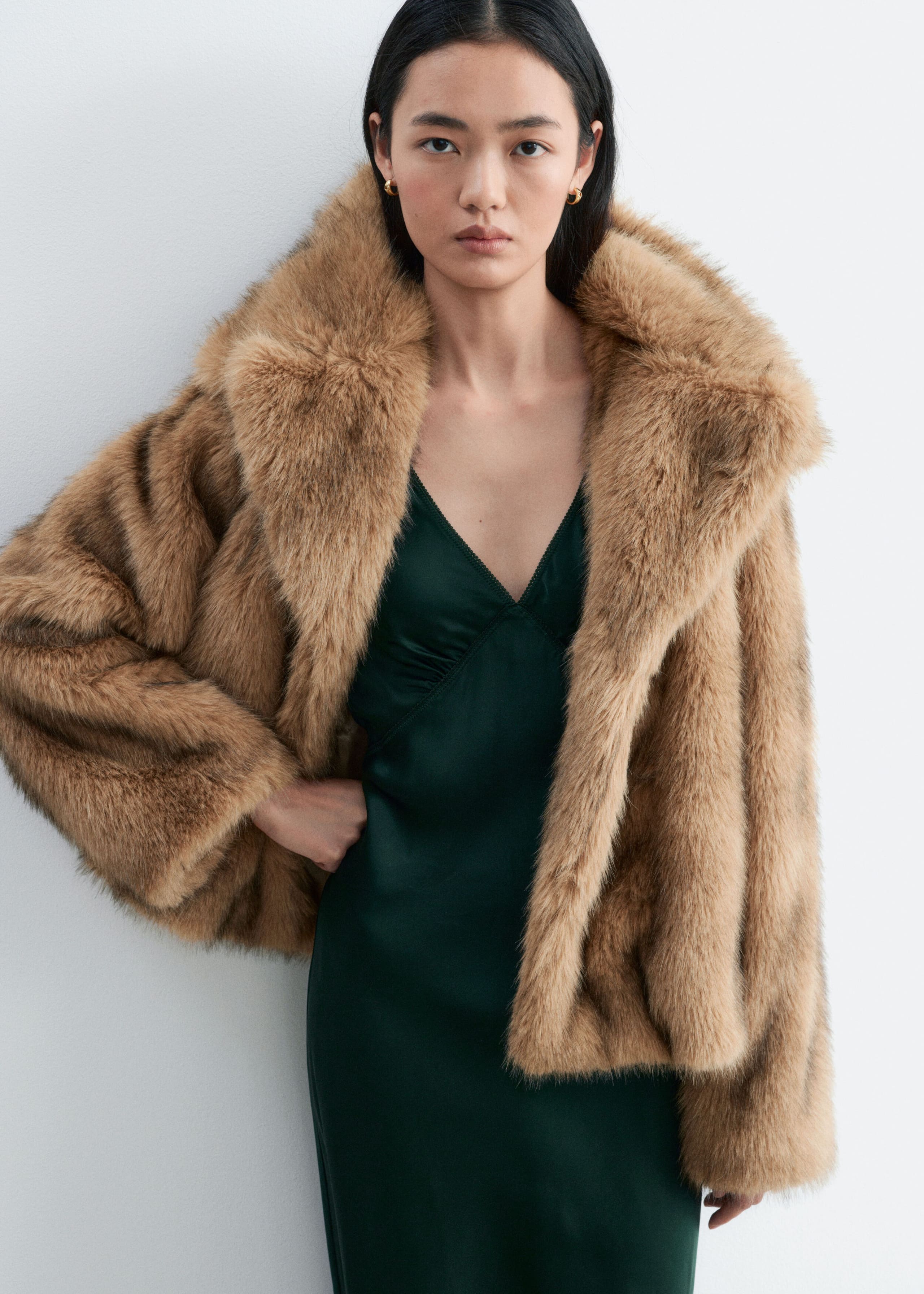 The model is wearing a dark green slip dress with a V-neckline and a softly gathered bust. The dress is styled under an open, oversized brown faux fur jacket.