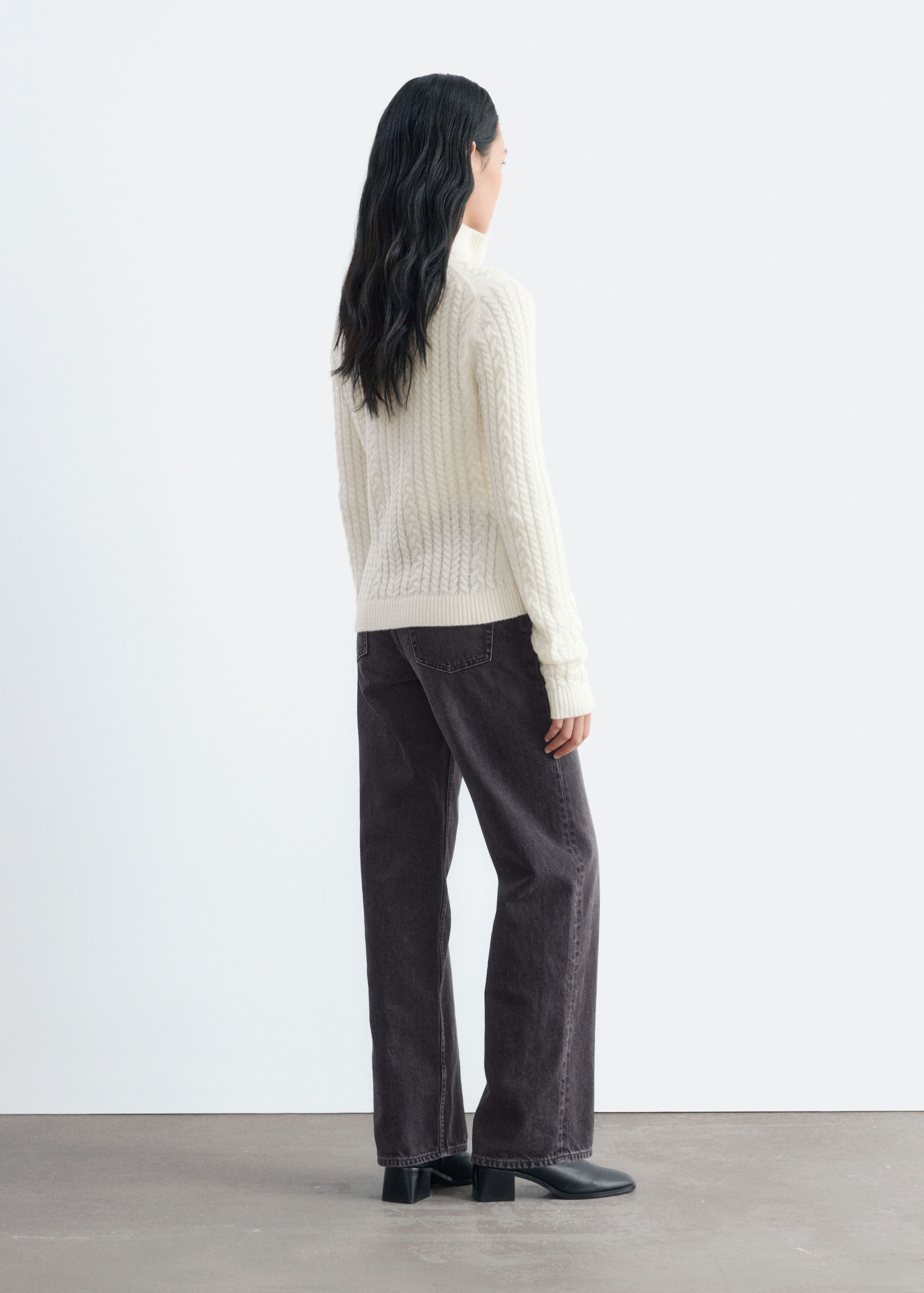 A cream-coloured, cable-knit cardigan with a high ribbed collar is being worn by the model, who is standing and facing away from the camera. The slim-fit cardigan is paired with dark grey wide-leg jeans and black leather boots.