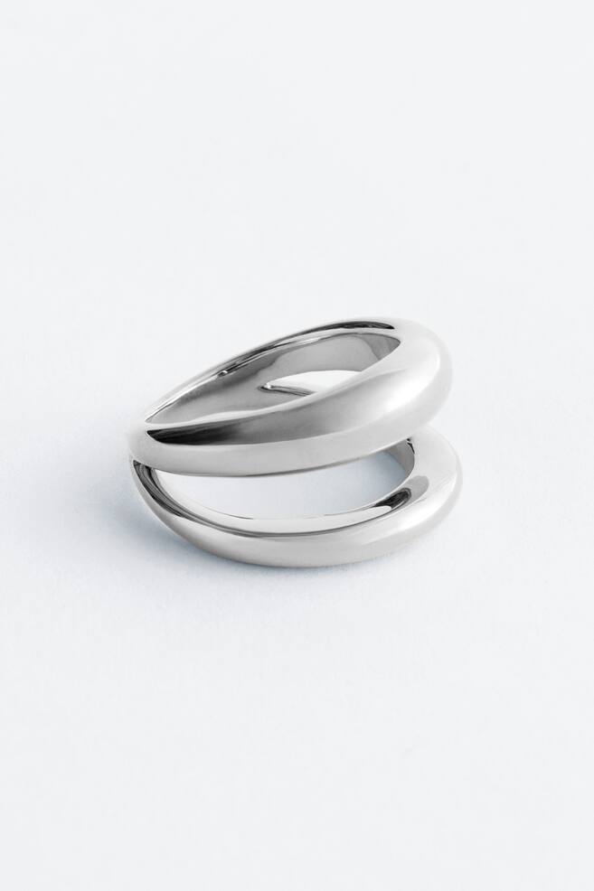 De ring toont een gepolijst zilverkleurig ontwerp met dubbele band, waarbij twee gladde, afgeronde banden zich krommen en scheiden om een gelaagde structuur te vormen. Hij is zo gepositioneerd dat zijn driedimensionale vorm wordt benadrukt. - 1