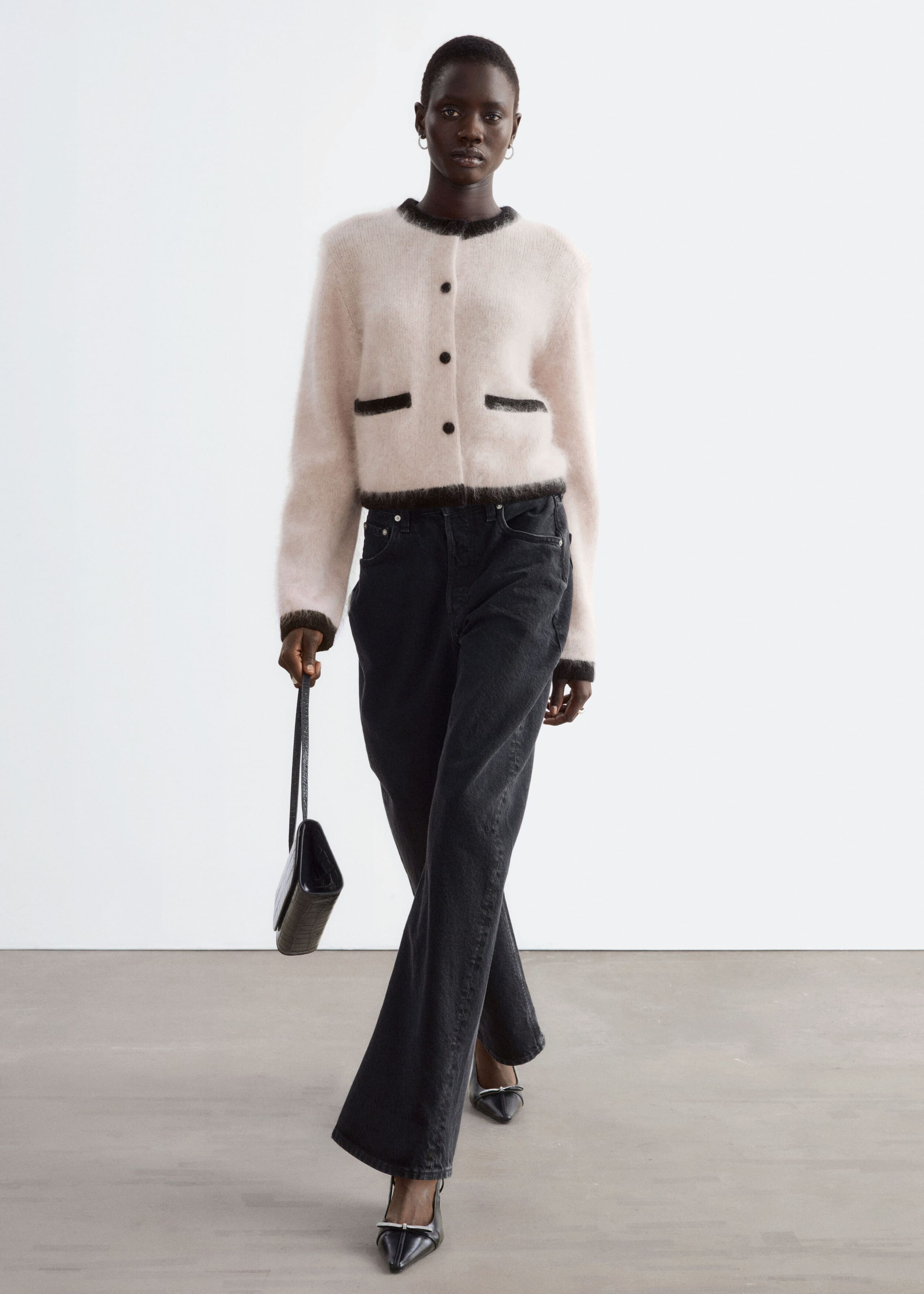 A light pink fuzzy knit cardigan with black contrast trim on the collar, cuffs, hem, and faux pocket details is worn by the model. The cardigan is styled with black wide-leg jeans, and the model is holding a small black handbag.