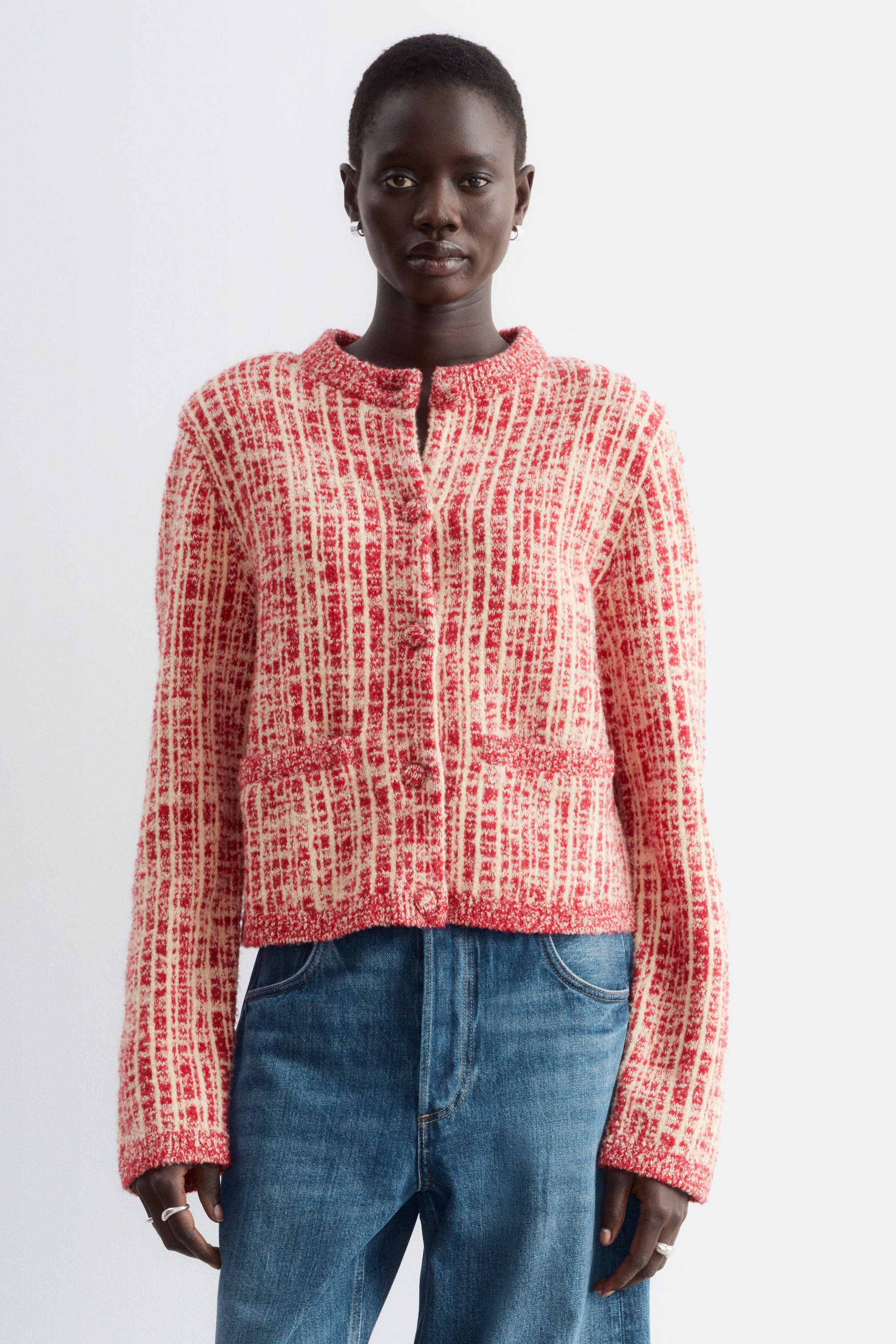 Grösseres Bild ansehen: Ein rot- und cremefarbenes Strick-Cardigan mit strukturierter Schulterpartie, Rippabschlüssen und mit Stoff bezogenen Frontknöpfen wird vom Model getragen. Das Cardigan ist zugeknöpft gestylt und mit blauen Denim-Jeans kombiniert.