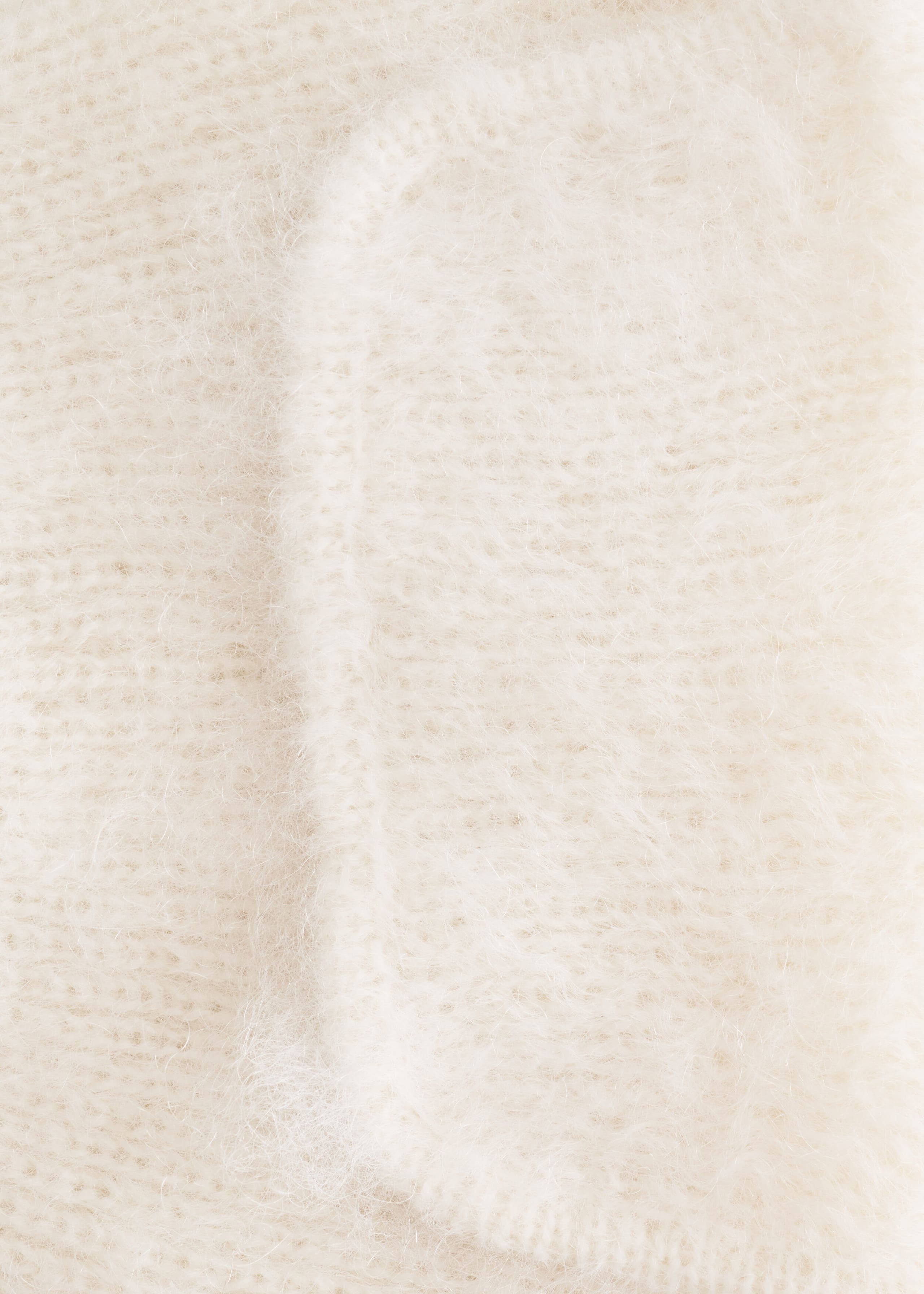 A close-up view of the cream-colored knit trapper hat showcases the soft, fuzzy texture of its wool and mohair blend. The shot highlights the rounded edge of an earflap stitched onto the main body, emphasizing the brushed, fibrous finish.