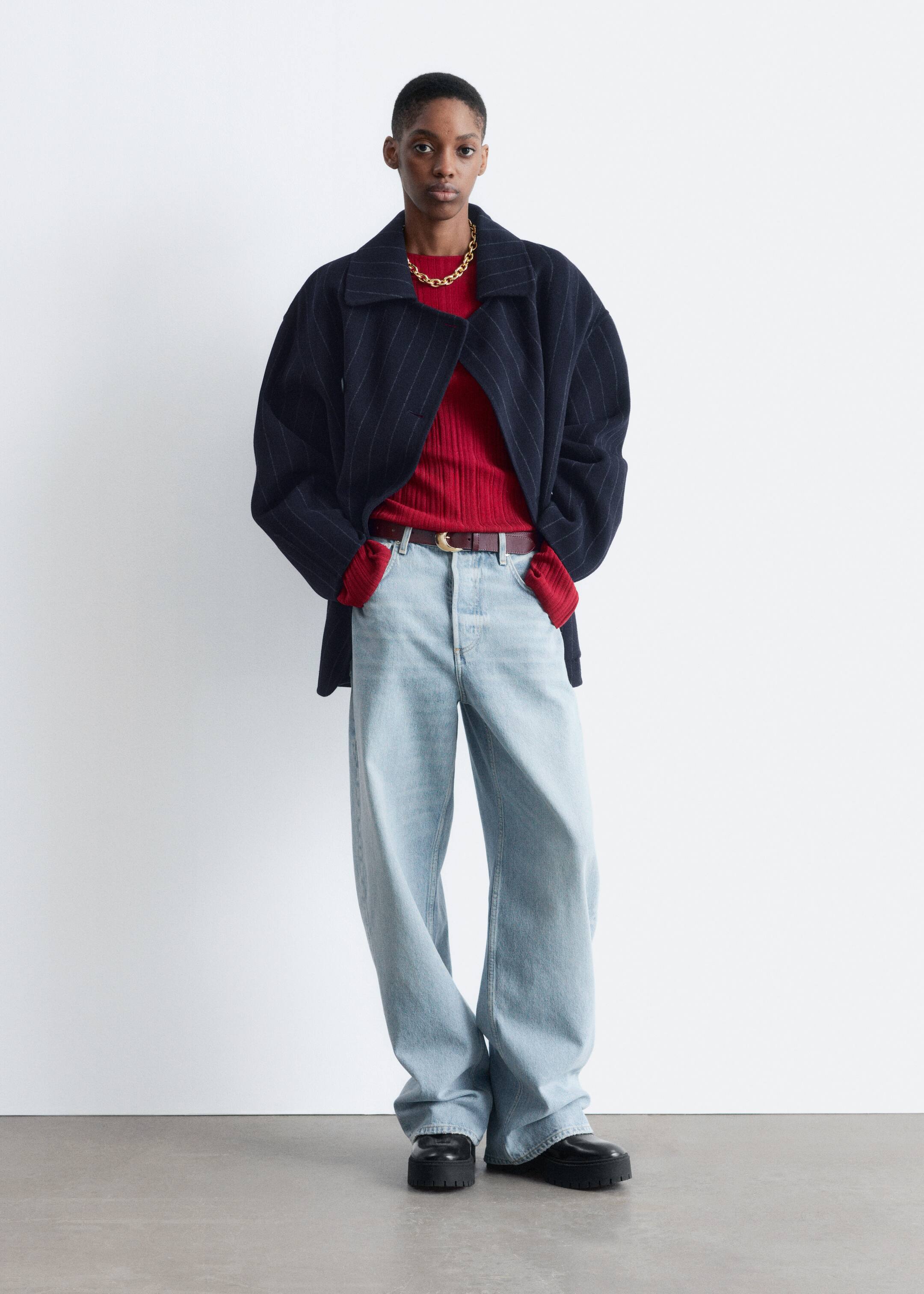 View larger image: A model is standing front-facing, wearing a navy pinstripe oversized jacket with wide lapels, layered over a red ribbed long-sleeved top and a gold-colored chain necklace. They are styling this with light wash, wide-leg jeans, a dark brown belt with a gold-toned buckle, and black chunky platform shoes.