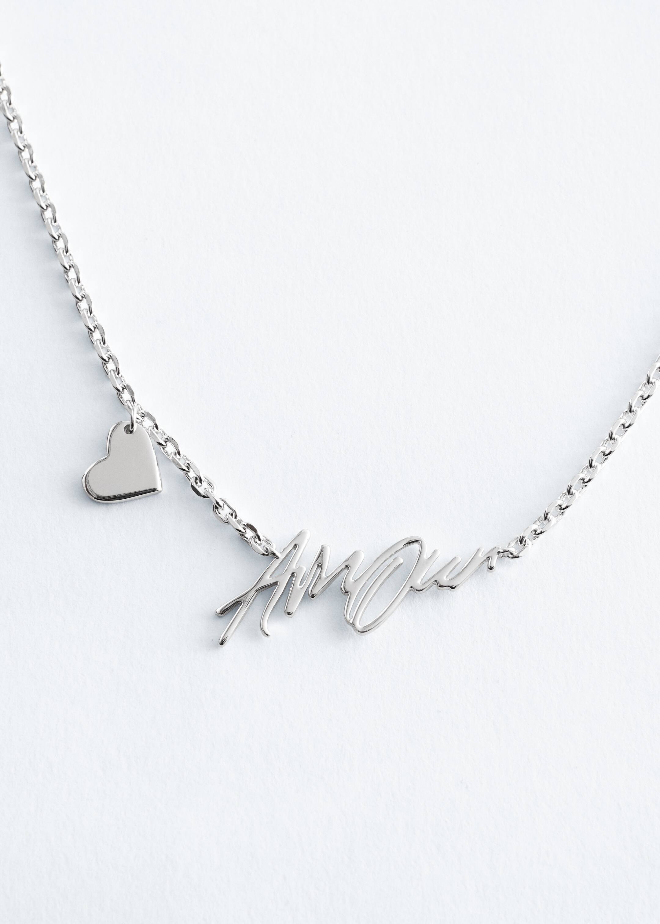 Visa större bild: Ett halsband i silvertonad metall har en polerad platt länkad kedja. Ett hängsmycke med kursiv text som stavar "Amour" är placerat i mitten fram, och en liten, polerad hjärtberlock hänger från kedjan till vänster.