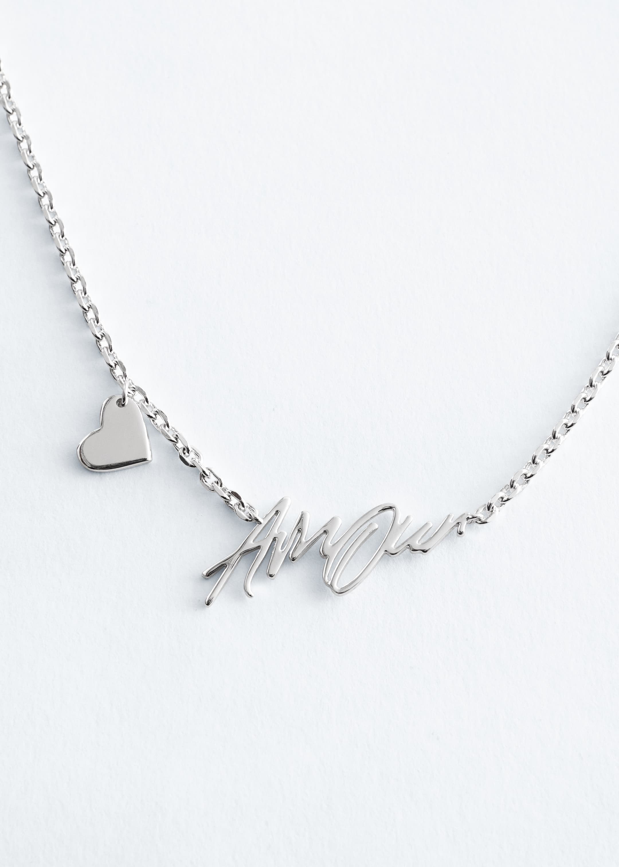 Grotere afbeelding bekijken: Een zilverkleurige metalen ketting heeft een gepolijste platte schakelketting. Een hanger met cursieve tekst die "Amour" spelt, is in het midden aan de voorkant geplaatst, en een kleine, gepolijste hartvormige bedel bungelt aan de linkerkant van de ketting.