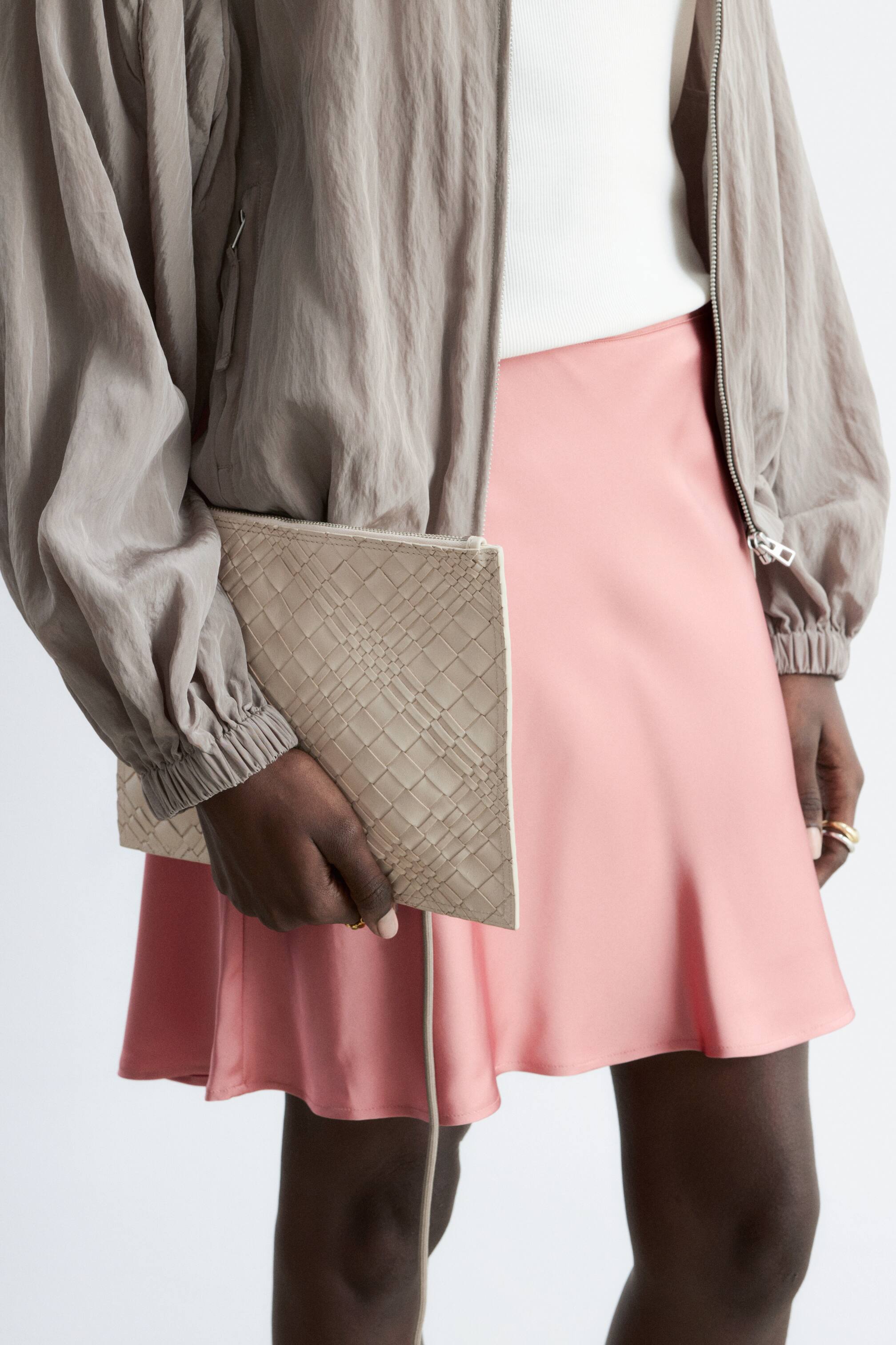 Преглед на по-голямо изображение: Плетен кожен портфейл с лека бежова текстура и горно закопчаване с цип, държан от модела. Тънка каишка виси от портфейла.