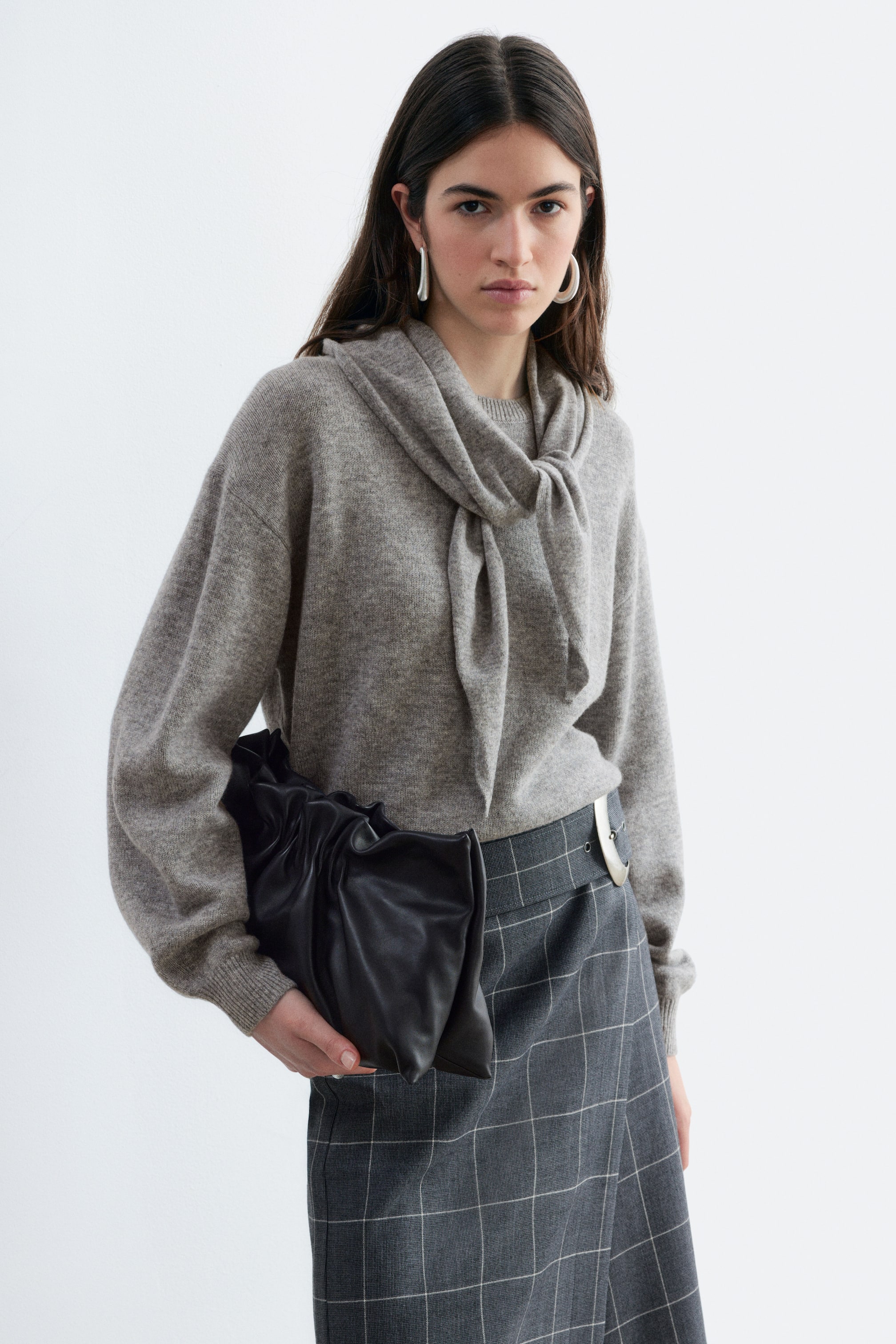 Visualizza l'immagine ingrandita: Un maglione in lana merino grigio mélange, dalla vestibilità rilassata, con polsini a coste e un dettaglio sciarpa integrato annodato morbidamente al collo, indossato dalla modella. Il maglione è abbinato infilato in una gonna scozzese grigio scuro, mentre la modella tiene in mano una pochette in pelle nera arricciata.