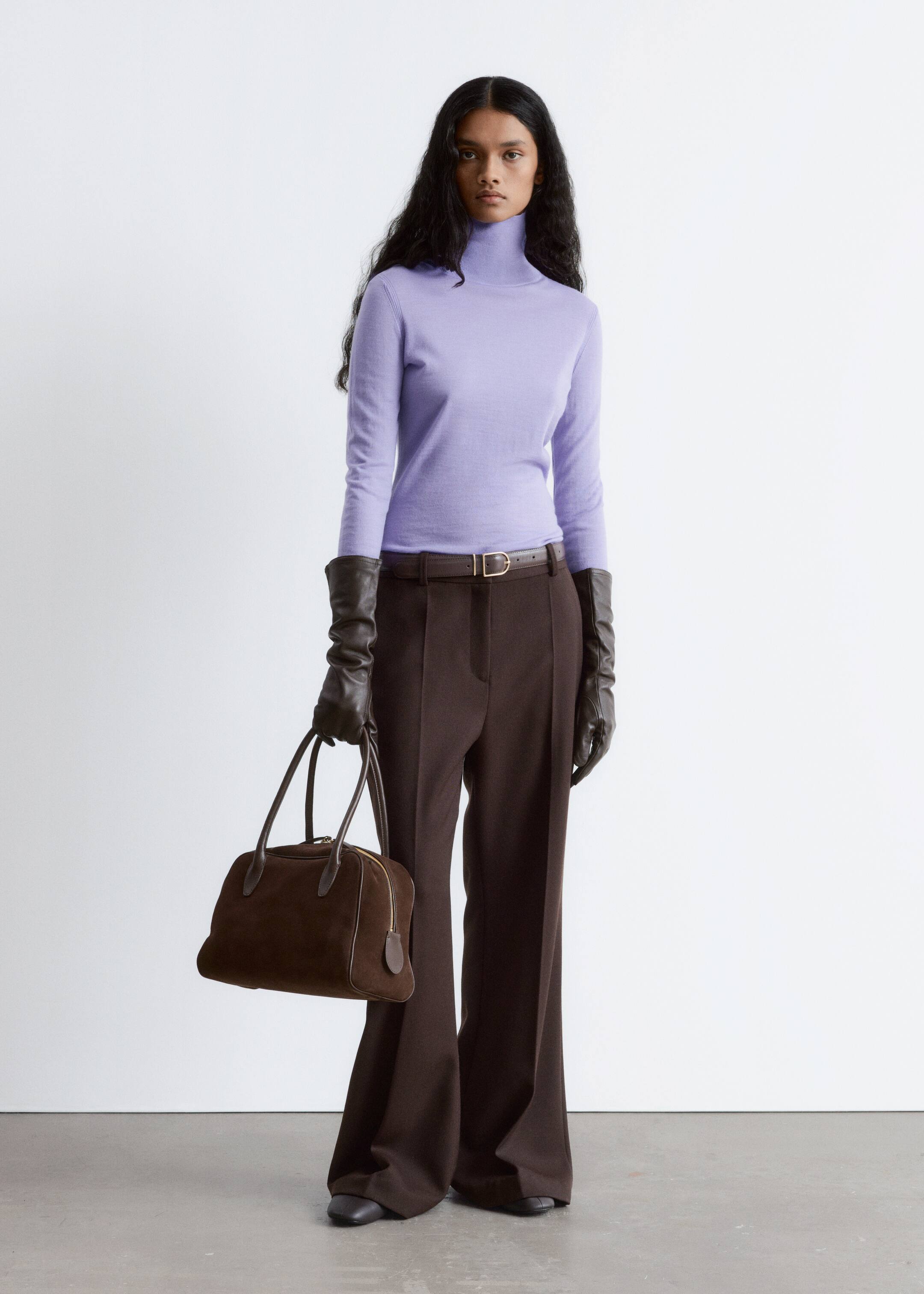 Grotere afbeelding bekijken: Een aansluitende lavendelkleurige coltrui, met een hoge hals en lange mouwen, wordt gepresenteerd. Het wordt gestyled met hoog getailleerde, wijde donkerbruine broek, aangesnoerd door een dunne donkerbruine riem met een goudkleurige gesp. Lange donkerbruine leren handschoenen worden gedragen, en een gestructureerde donkerbruine suède en leren handtas wordt vastgehouden.