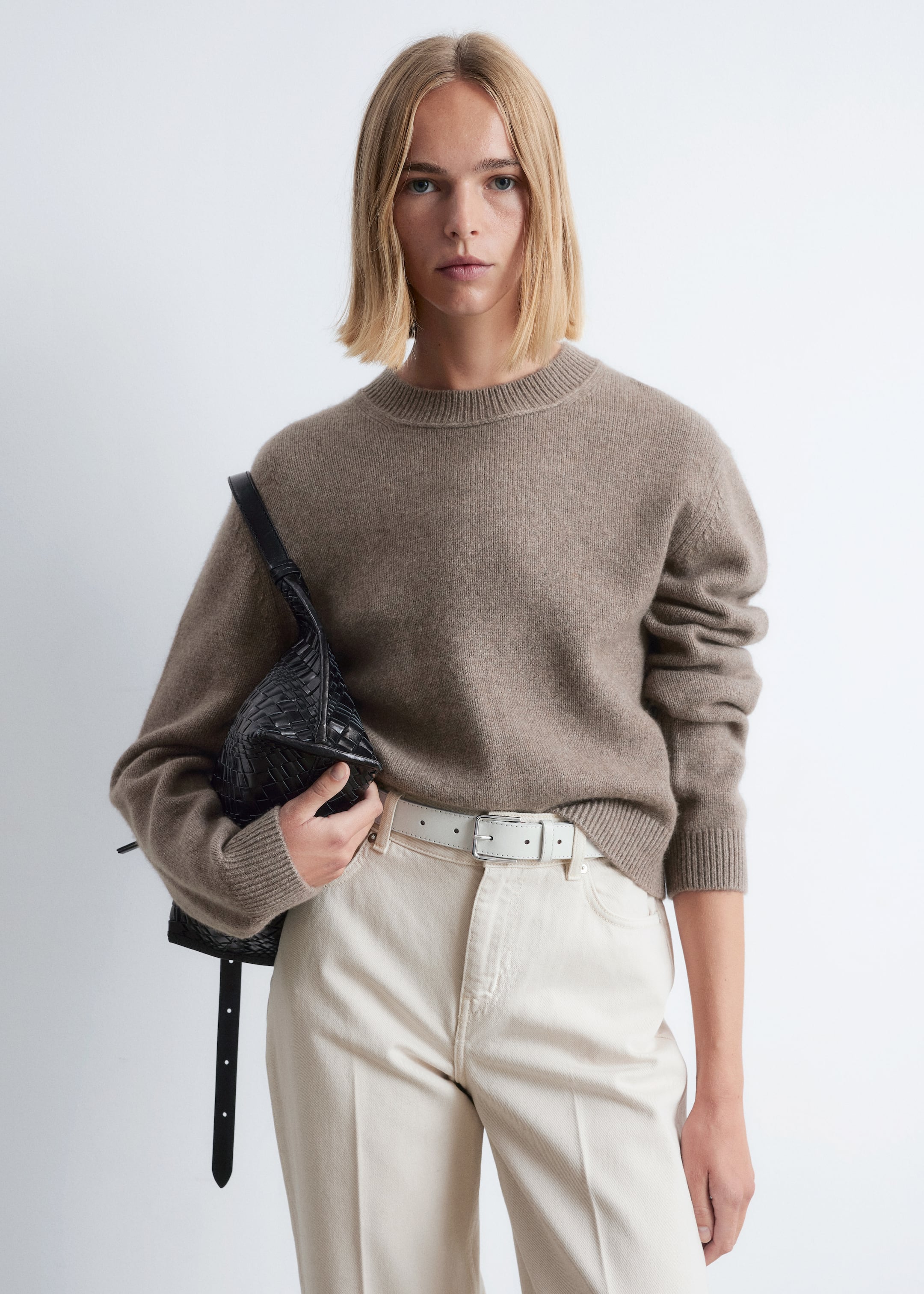 Ingrandisci l'immagine: Un corto maglione lavorato a maglia color talpa con bordi a coste e spalle scese è indossato da una modella. Il maglione si appoggia sopra pantaloni di colore chiaro, e la modella tiene una borsa intrecciata nera contro il suo fianco.