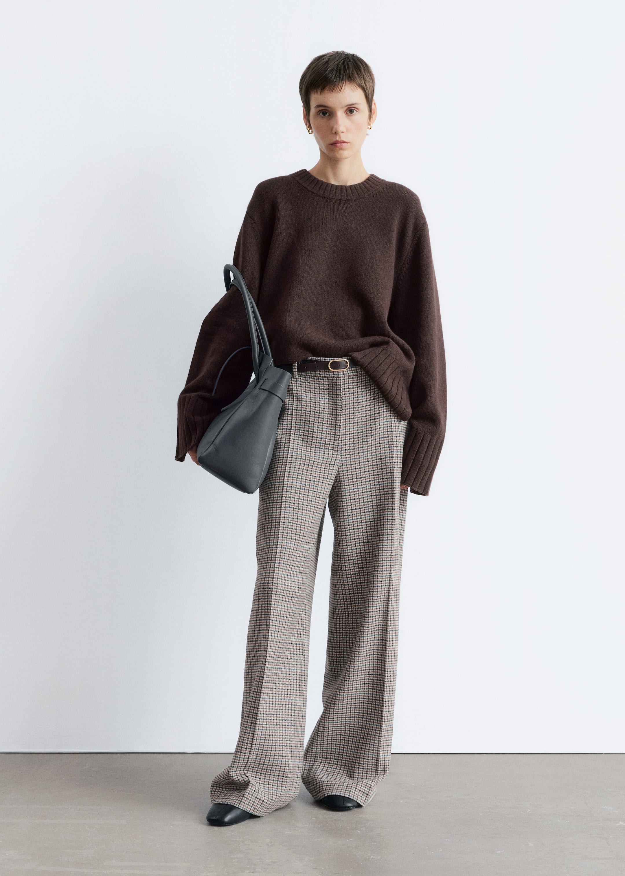 Agrandir l'image: Un pull à col rond marron foncé avec des poignets côtelés est porté, rentré de manière lâche dans un pantalon à carreaux à jambes larges. Le pantalon est maintenu par une fine ceinture avec une boucle ovale, et un sac en cuir souple gris foncé est tenu. Des chaussures plates noires sont également visibles.