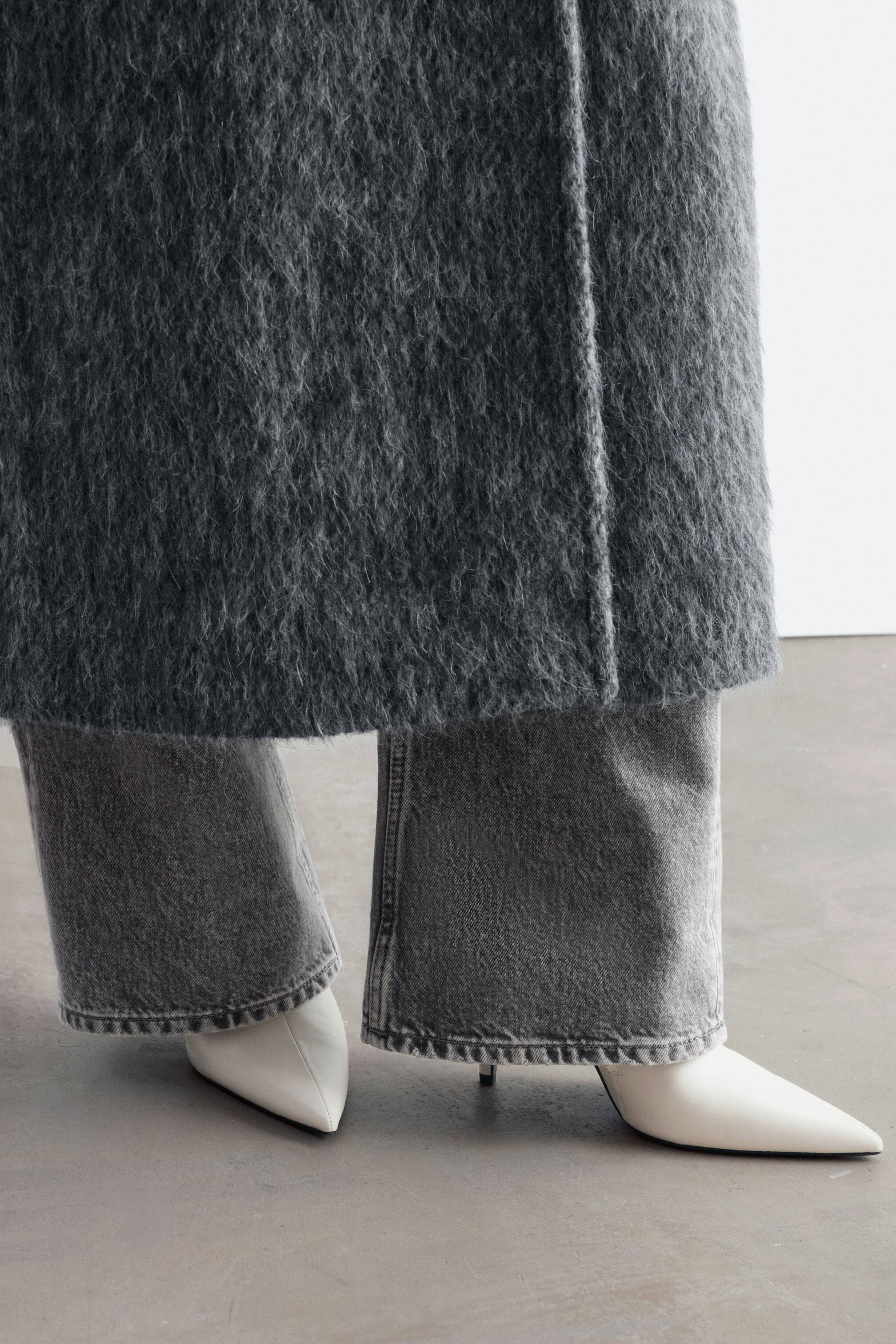 Grotere afbeelding bekijken: Witte leren enkellaarzen met een spitse neus en hoge stilettohak worden gedragen door het model. De laarzen zijn gestyled onder een wijde grijze jeans en een lange, getextureerde grijze mantel.