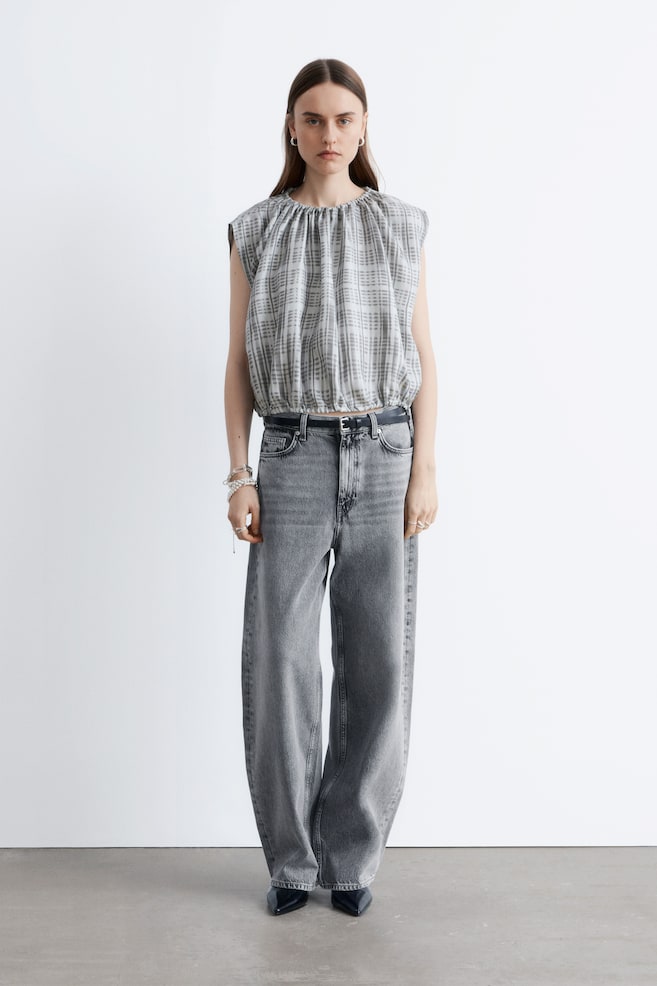 Un haut sans manches arbore un motif à carreaux gris et blanc, avec un tissu froncé au niveau de l'encolure ronde et un ourlet élastiqué, formant une coupe blouson. Il est associé à un jean large taille haute gris délavé qui présente une silhouette décontractée, et une ceinture noire est passée dans les passants. - 1