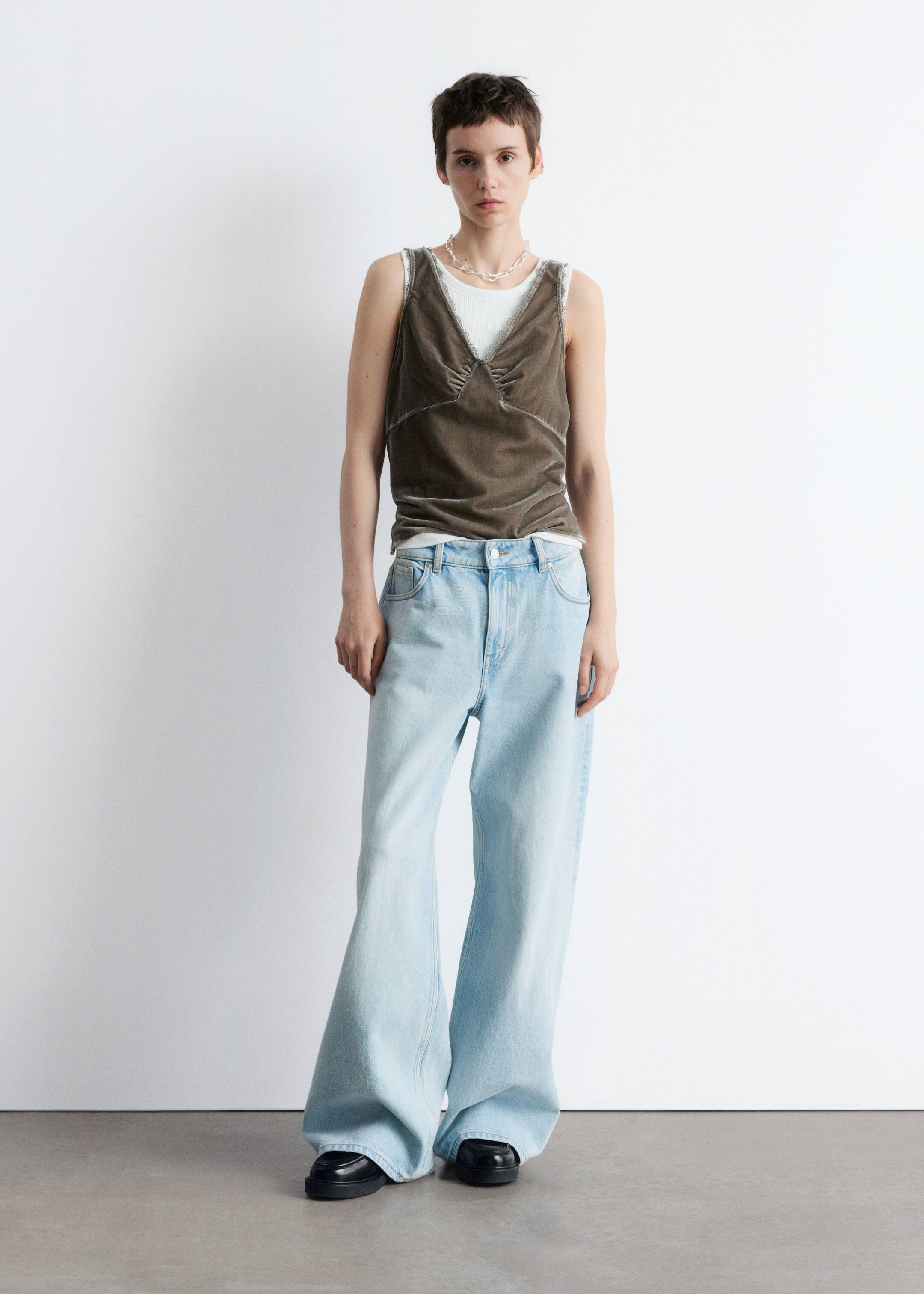 Größeres Bild anzeigen: Ein Outfit wird direkt präsentiert, mit einem dunkel olivgrünen Samt-Camisole mit V-Ausschnitt, gerafften Details und Spitzenbesatz entlang des Ausschnitts und der Armlöcher, über einem weißen gerippten Tanktop. Helle, weit geschnittene Jeans werden getragen, die über schwarzen, klobigen Plateauschuhen liegen, ergänzt durch eine klare, klobige Kettenhalskette.