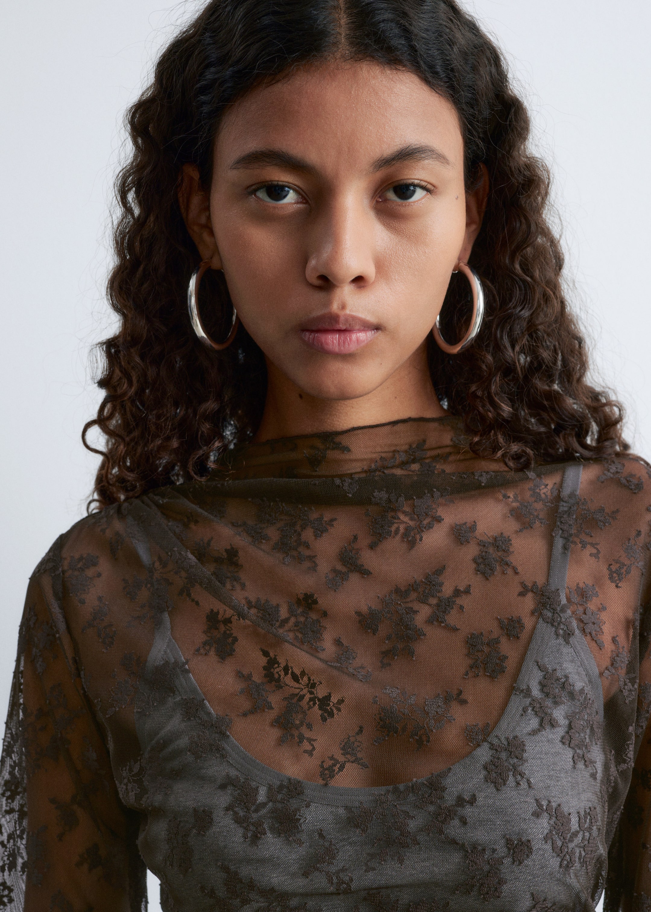 Agrandir l'image: Un haut sombre et transparent est porté, présenté de face et doté d'un col montant drapé avec un motif floral sombre brodé sur toute la surface. Il se superpose à un caraco gris uni, mettant en valeur son design translucide.