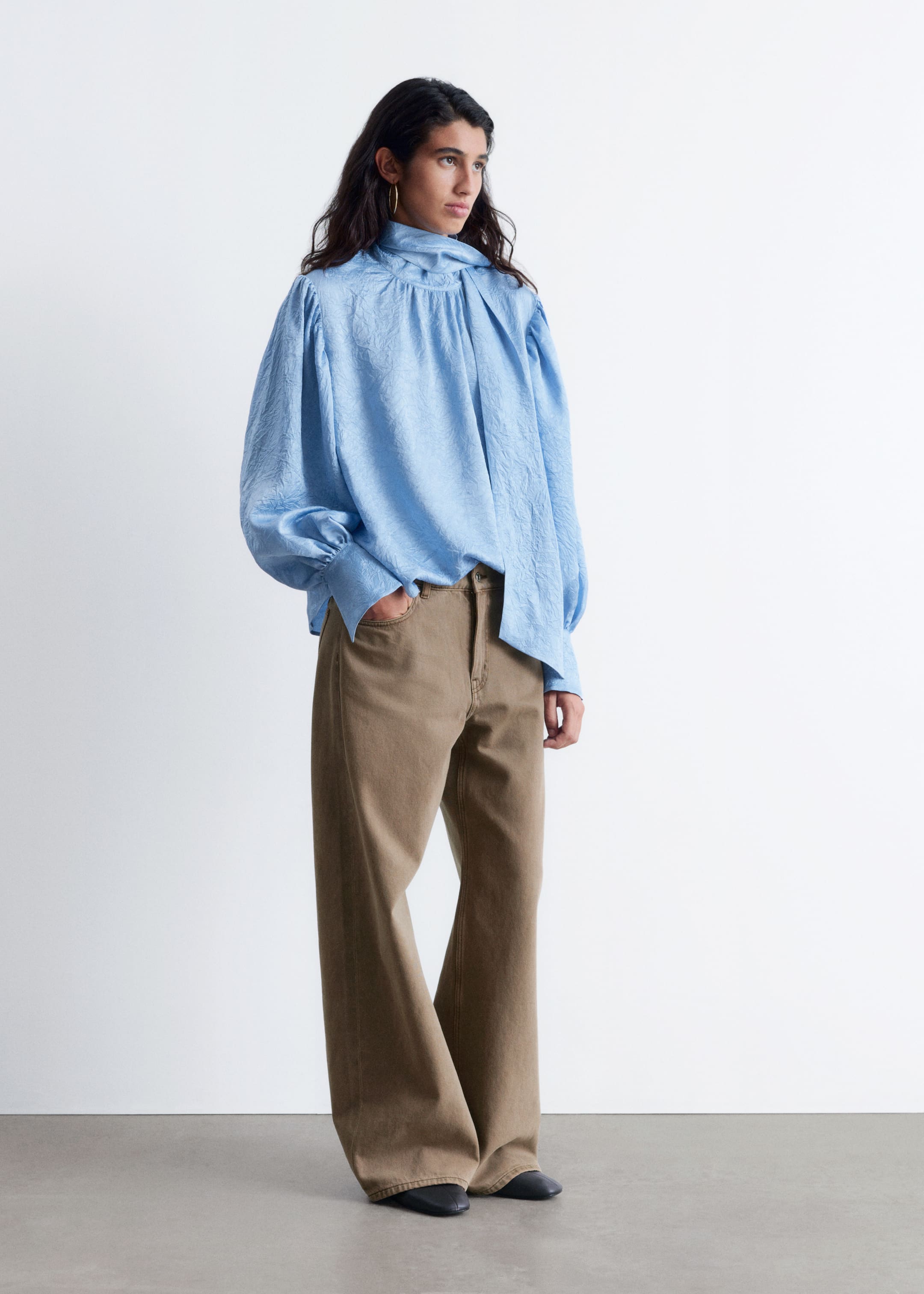 Grotere afbeelding bekijken: Een lichtblauwe blouse met een subtiele kreukeltextuur wordt gepresenteerd, met een hoge, geplooide halslijn en een gedrapeerd strikdetail. De blouse heeft volumineuze lange mouwen die zijn geplooid bij de schouders en manchetten, en is gedeeltelijk ingestopt in hoog getailleerde, wijde kaki bruine broek.