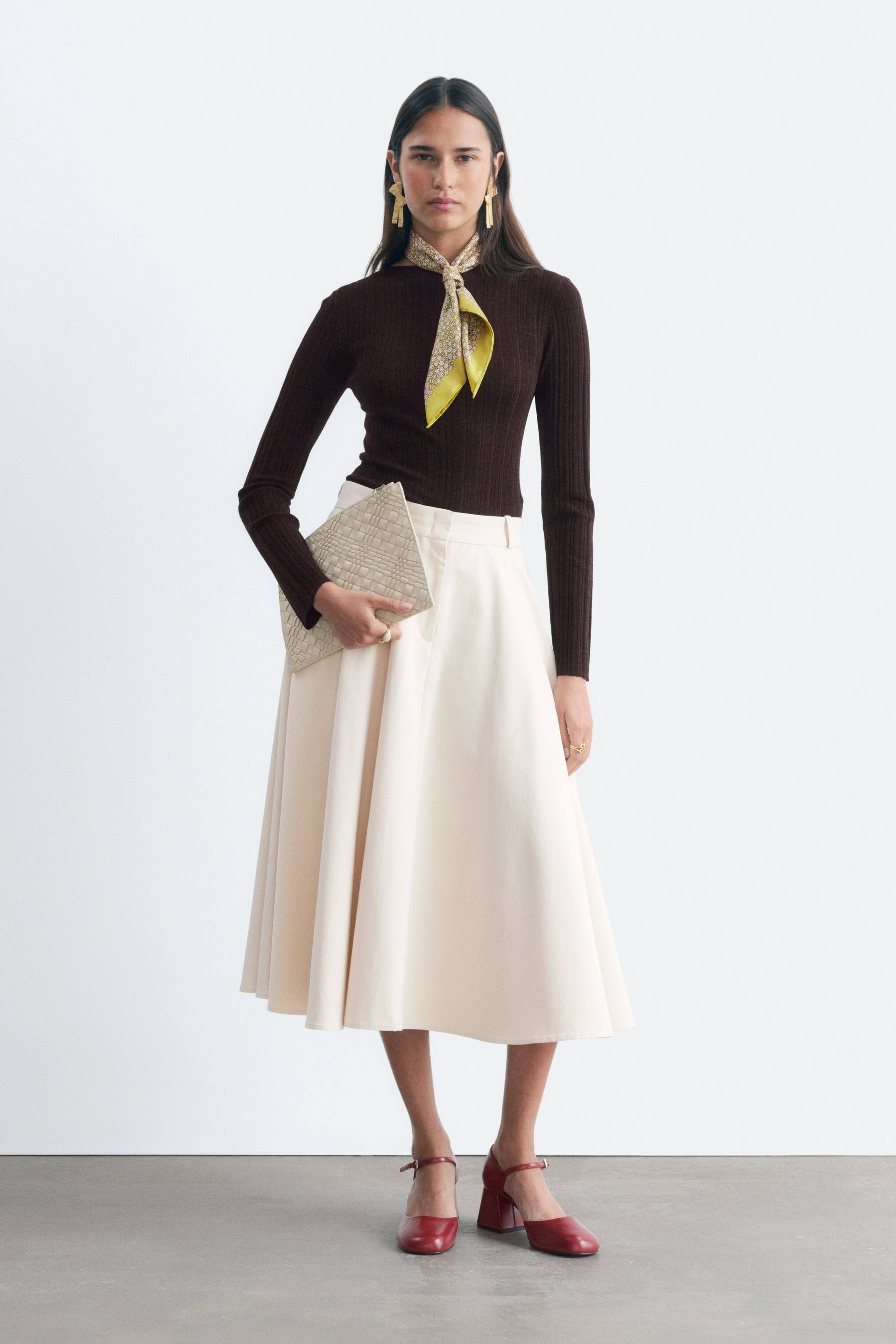 View larger image: The model is wearing a cream circle-cut midi skirt, which features a zip fly, belt loops, and slanted side pockets. The skirt is paired with a dark brown ribbed top.