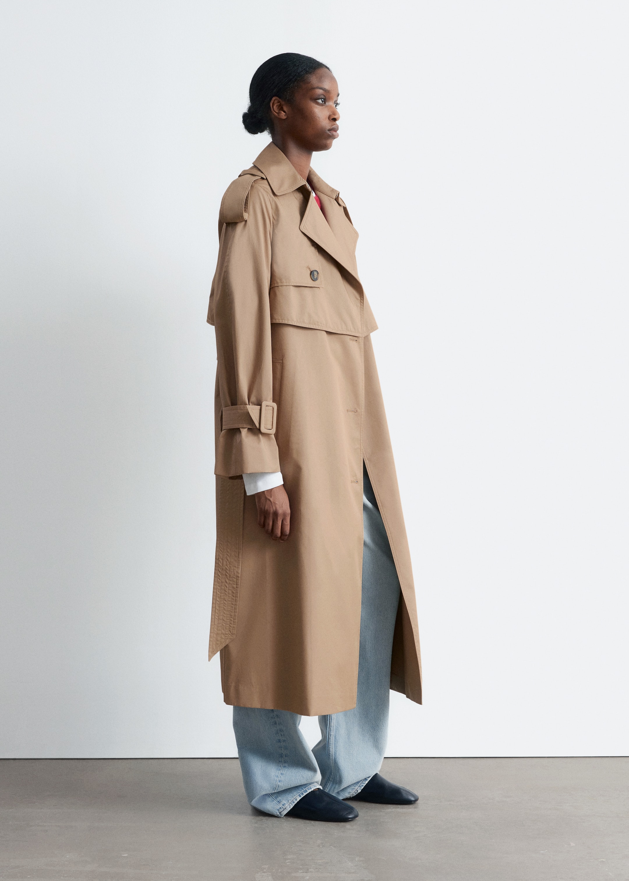 Visualizza immagine più grande: Un lungo trench coat doppiopetto color sabbia viene presentato di profilo. Presenta ampi revers, bottoni visibili sul pannello frontale sovrapposto, spalline sulle spalle, uno scudo antipioggia sulla parte superiore della schiena e cinturini con fibbia ai polsini. Il cappotto cade morbido, estendendosi sotto il ginocchio, ed è stratificato su una camicia bianca e jeans a gamba larga azzurri.