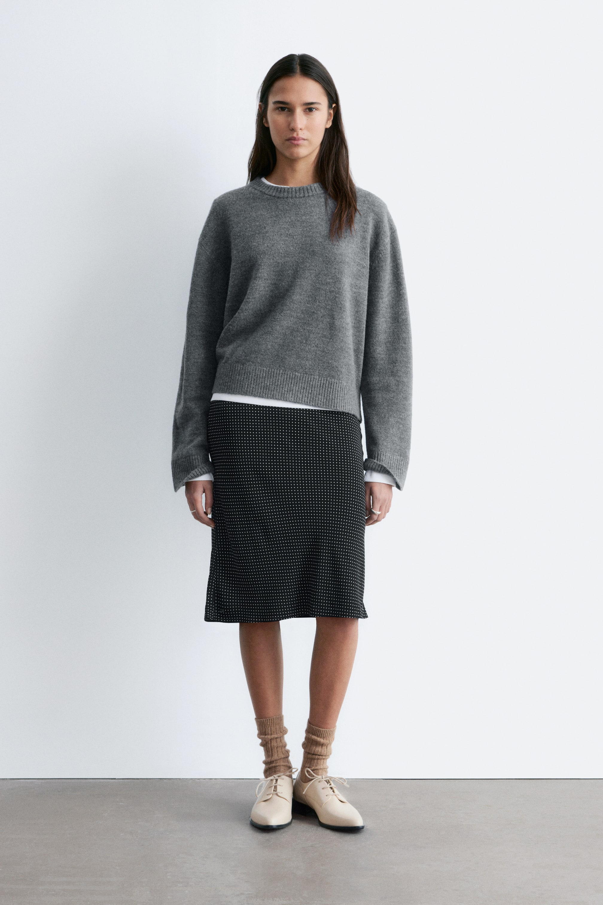 Преглед на по-голямо изображение: Сив пуловер с кръгло деколте е показан отпред, с релаксирана кройка, дълги ръкави, рипсени маншети и рипсен подгъв. Бяла риза се подава изпод подгъва и маншетите му.