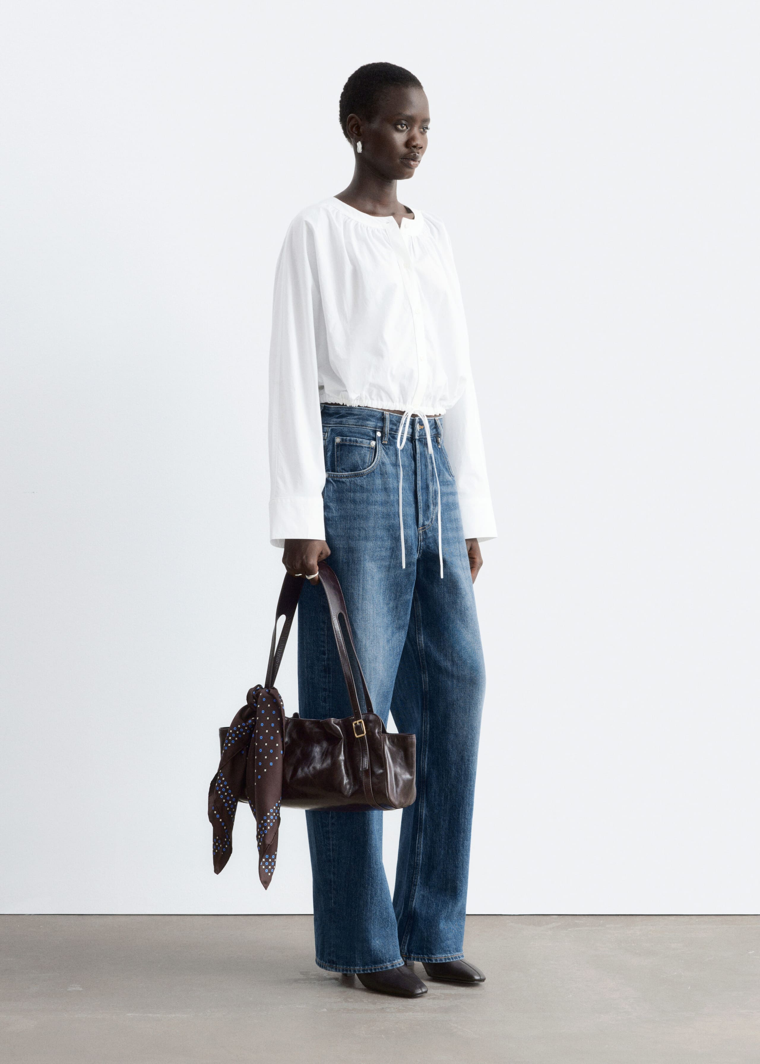 A white cropped cotton blouse featuring long sleeves, a gathered neckline, and a drawstring hem with hanging self-ties is being worn by the model. The blouse is styled with blue wide-leg jeans while the model holds a dark brown leather handbag.