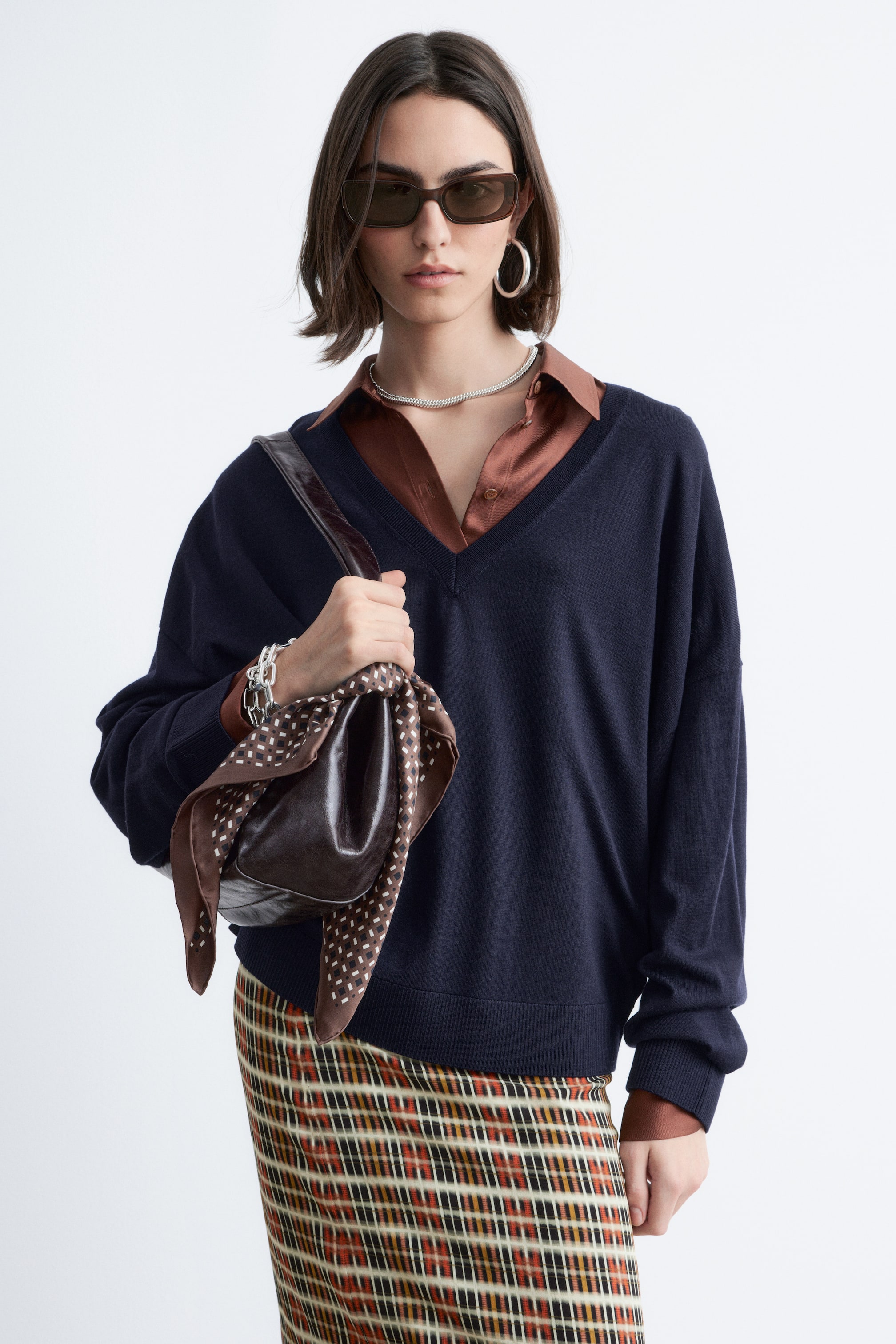 הצגת תמונה גדולה יותר: סוודר בצבע כחול כהה עם מחשוף V עמוק, שרוולים ארוכים וגזרה רפויה. הוא מציג חפתים וגימור תחתון מצולעים, ומונח בשכבות מעל חולצה עם צווארון חום הנראית במחשוף ובקצות השרוולים.