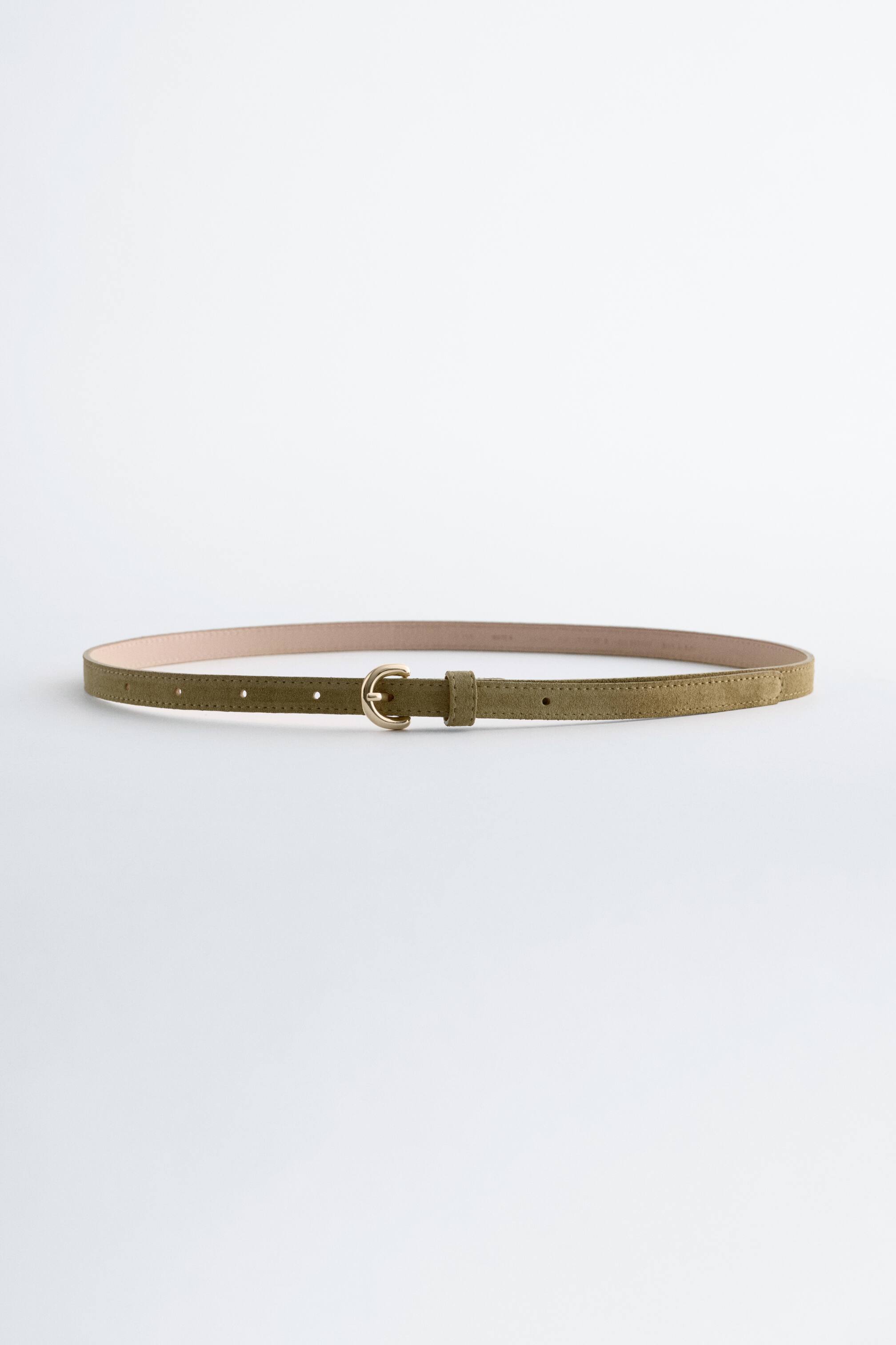 Agrandir l'image: Une ceinture en daim vert olive est posée à plat, présentant une fine lanière avec un dessous beige clair et une petite boucle de ton laiton. Elle met en avant des coutures visibles le long de ses bords et plusieurs trous d'ajustement.