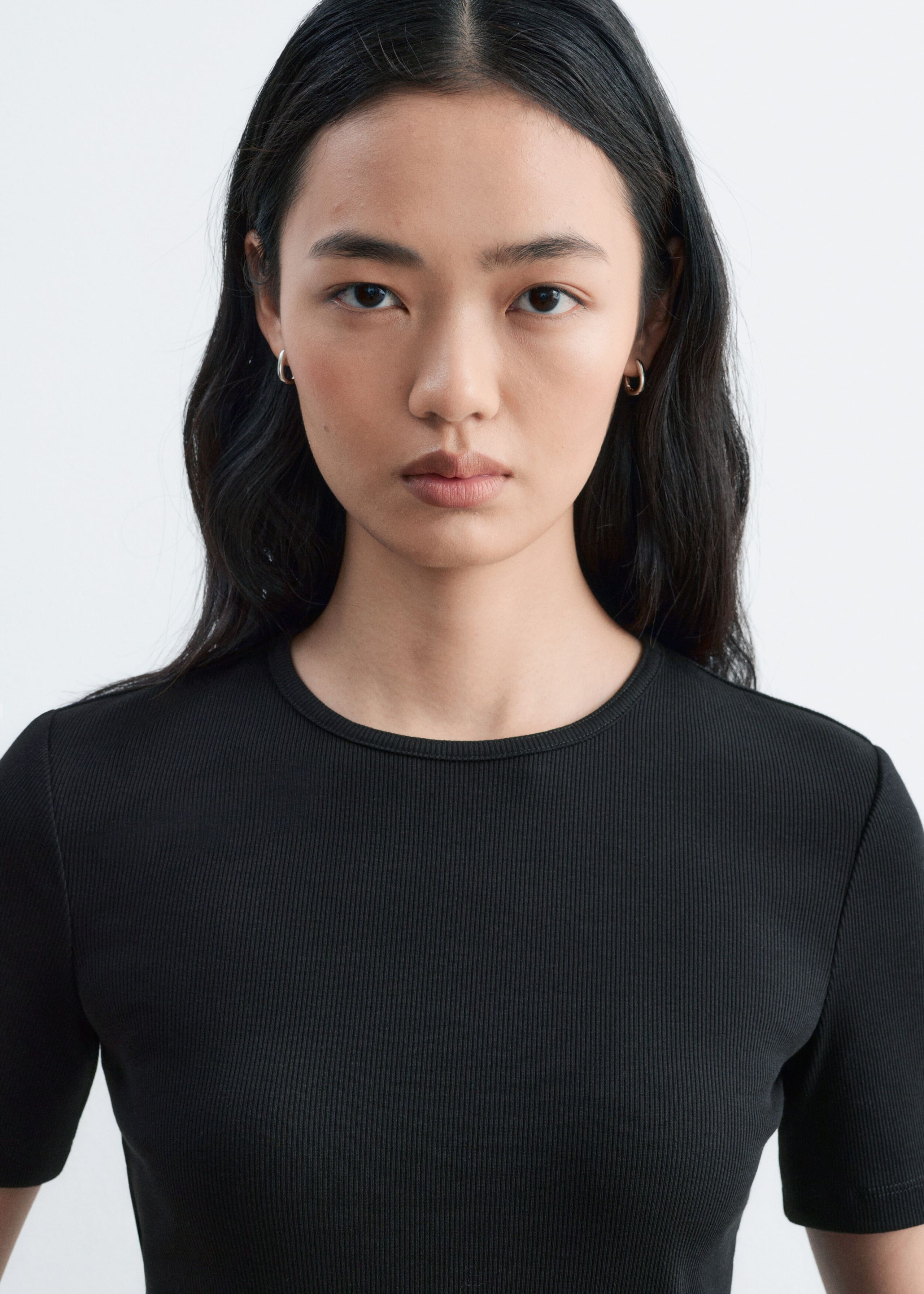 A black ribbed T-shirt featuring a round neckline and short sleeves is being worn by the model. This close-up view highlights the fine vertical ribbing and slim fit of the garment.