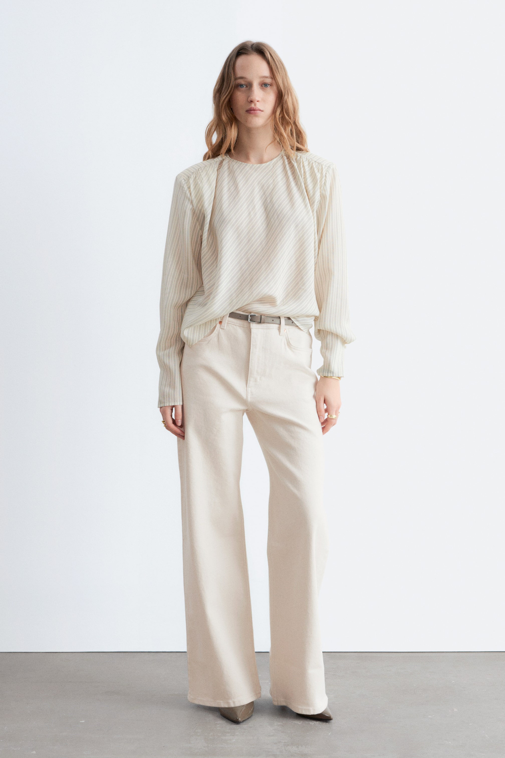 הצגת תמונה גדולה יותר: חולצה בצבע קרם עם פסים אלכסוניים עדינים, צווארון עגול וכתפיים מכווצות נלבשת כשהיא תחובה במכנסיים בצבע קרם בגזרה גבוהה ורחבה. חגורה דקה בצבע אפור מהדקת את המותן, ונעליים מחודדות בצבע אפור כהה משלימות את ההופעה.