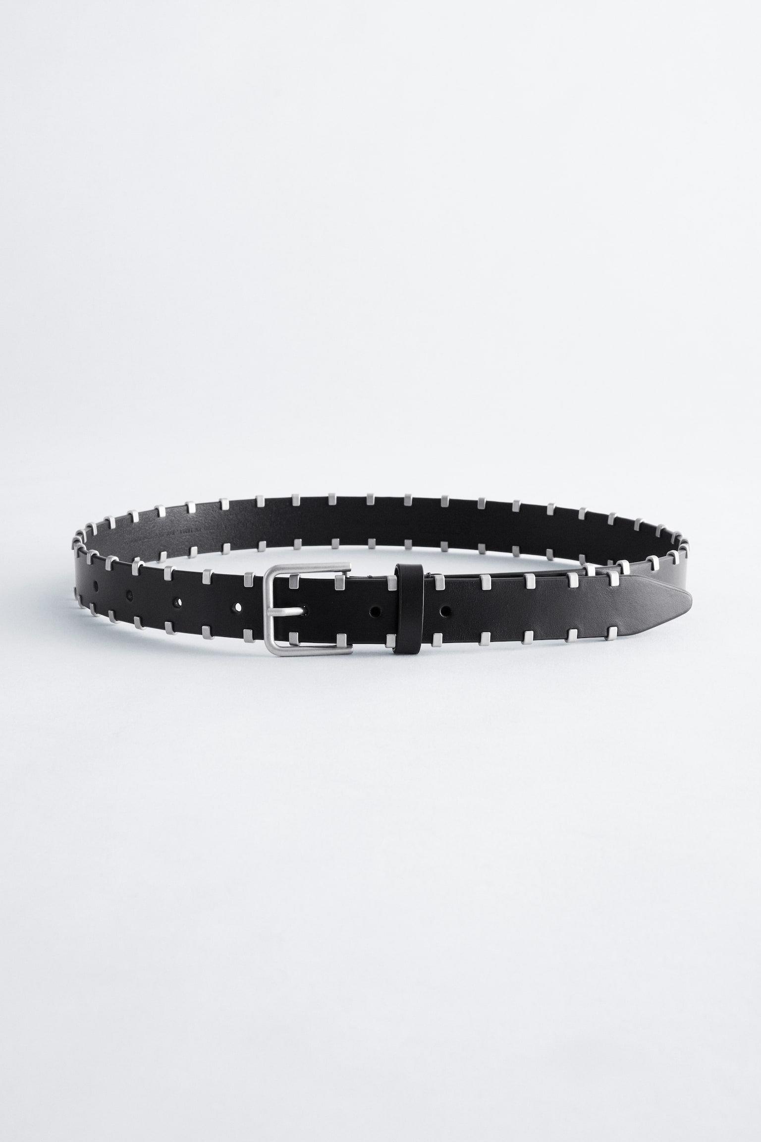 Een zwarte leren riem ligt in een losse lus naar voren gericht, met kleine, vierkante metalen studs in zilverkleur langs zowel de boven- als onderrand. De riem is afgewerkt met een rechthoekige zilverkleurige gesp, een bijpassende leren riemlus en voorgeponste gaatjes bij het taps toelopende uiteinde.