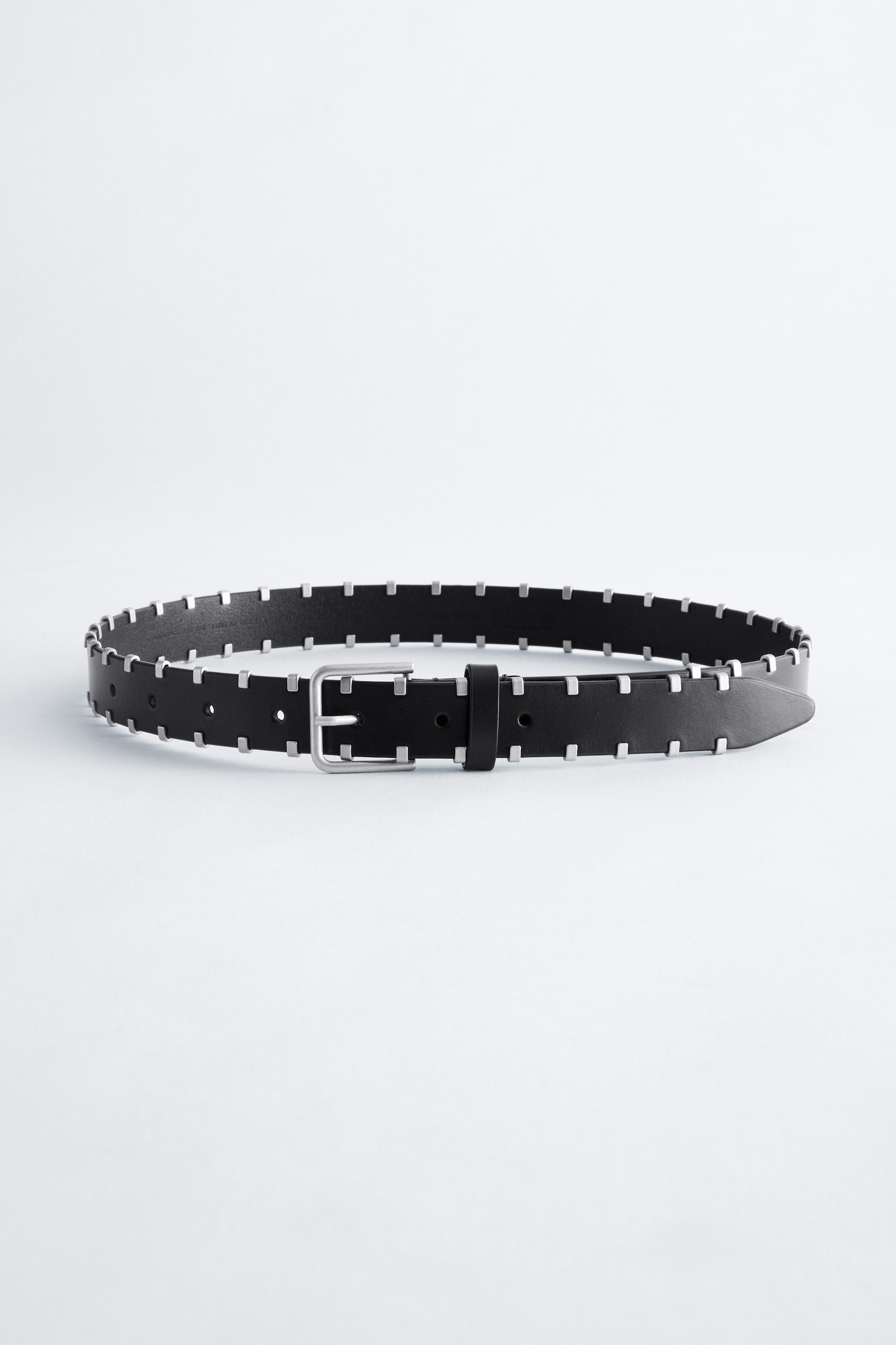 Grotere afbeelding bekijken: Een zwarte leren riem ligt in een losse lus naar voren gericht, met kleine, vierkante metalen studs in zilverkleur langs zowel de boven- als onderrand. De riem is afgewerkt met een rechthoekige zilverkleurige gesp, een bijpassende leren riemlus en voorgeponste gaatjes bij het taps toelopende uiteinde.