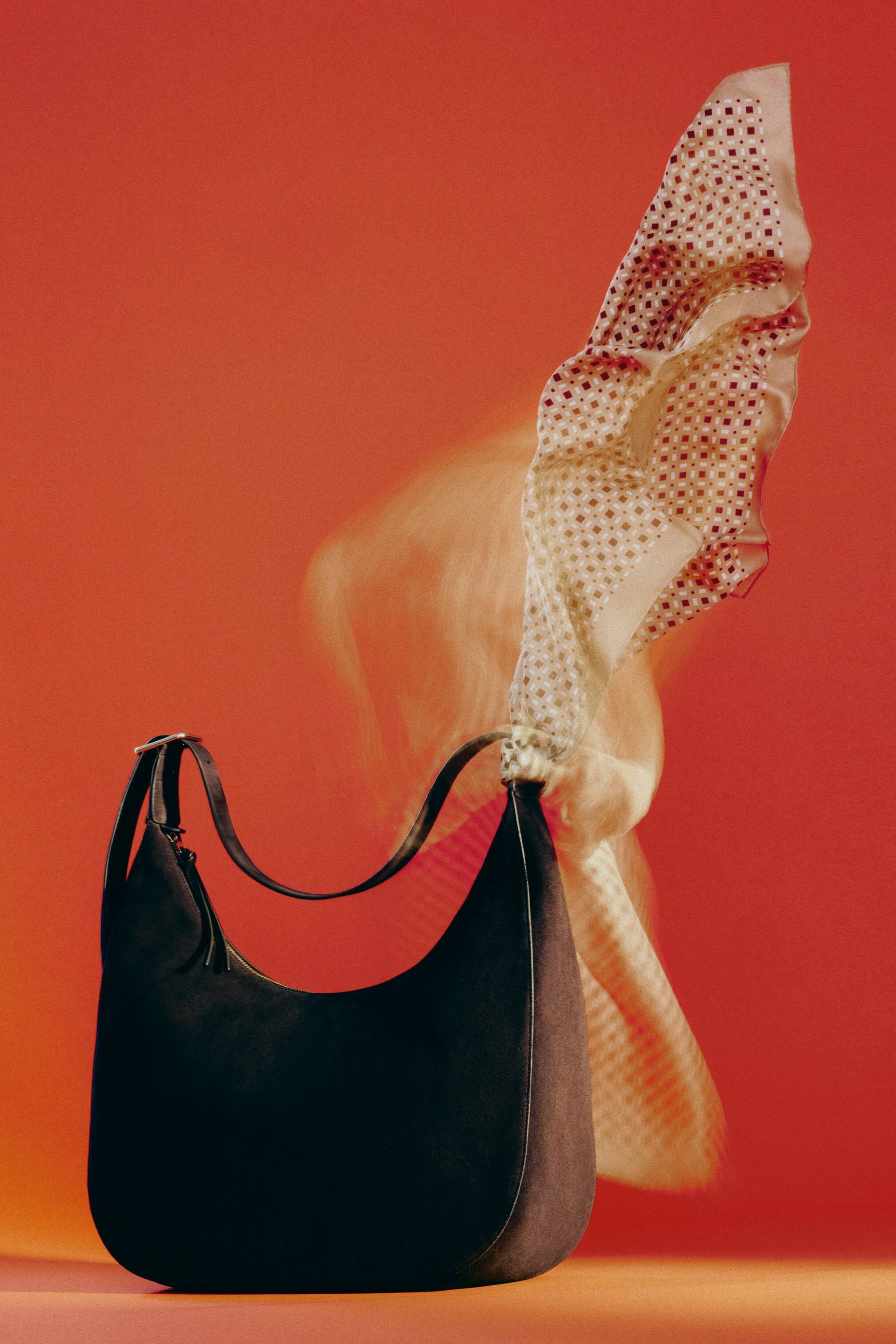 Agrandir l'image: Un sac hobo en forme de croissant, de couleur sombre et à texture mate avec des finitions lisses, est présenté sous un angle de face. Il est doté d'une fine bandoulière avec une boucle argentée et d'une fermeture éclair supérieure avec un tirette à pompon, tandis qu'un foulard à motifs de couleur claire est drapé sur sa bandoulière droite.