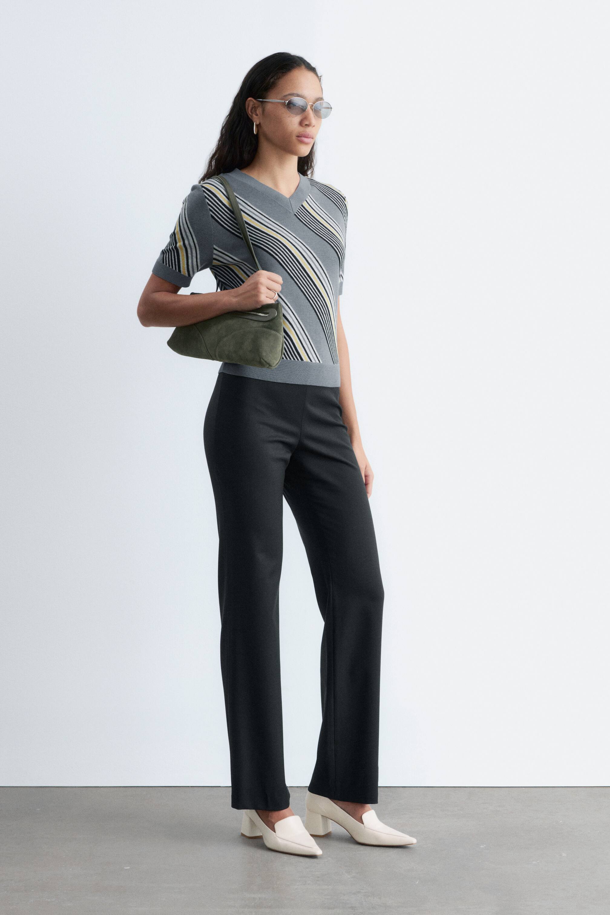 הצגת תמונה גדולה יותר: מכנסיים שחורים עם סילואטה נקייה ומינימליסטית ורגליים בגזרה ישרה נלבשים על ידי הדוגמנית, משולבים עם חולצה מפוספסת ונעליים בצבע בהיר.