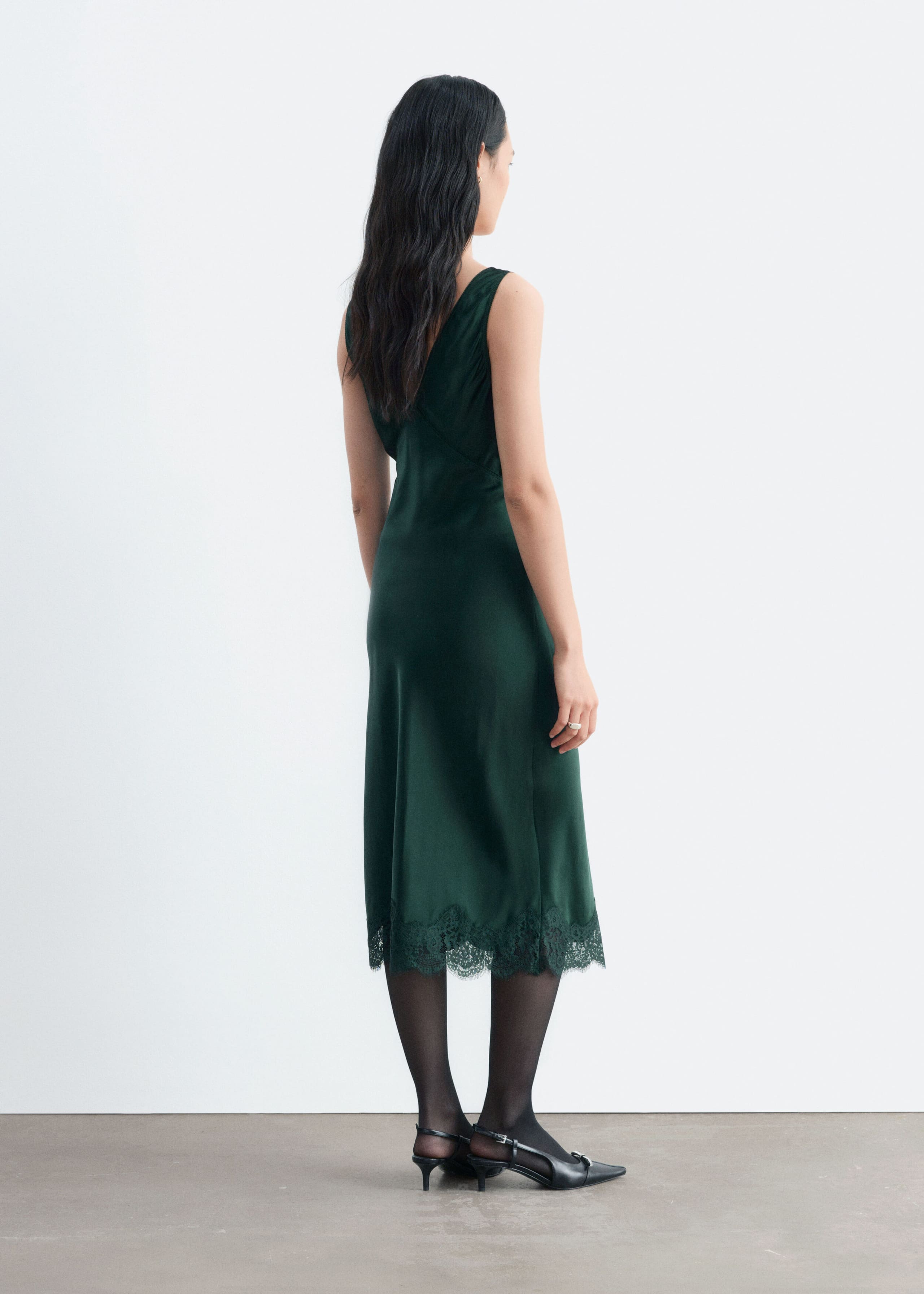 The model is wearing a dark green satin slip dress, showcasing its V-neck back, straight silhouette, and delicate scalloped lace hem. The dress is styled with black sheer tights and black pointed-toe slingback heels.