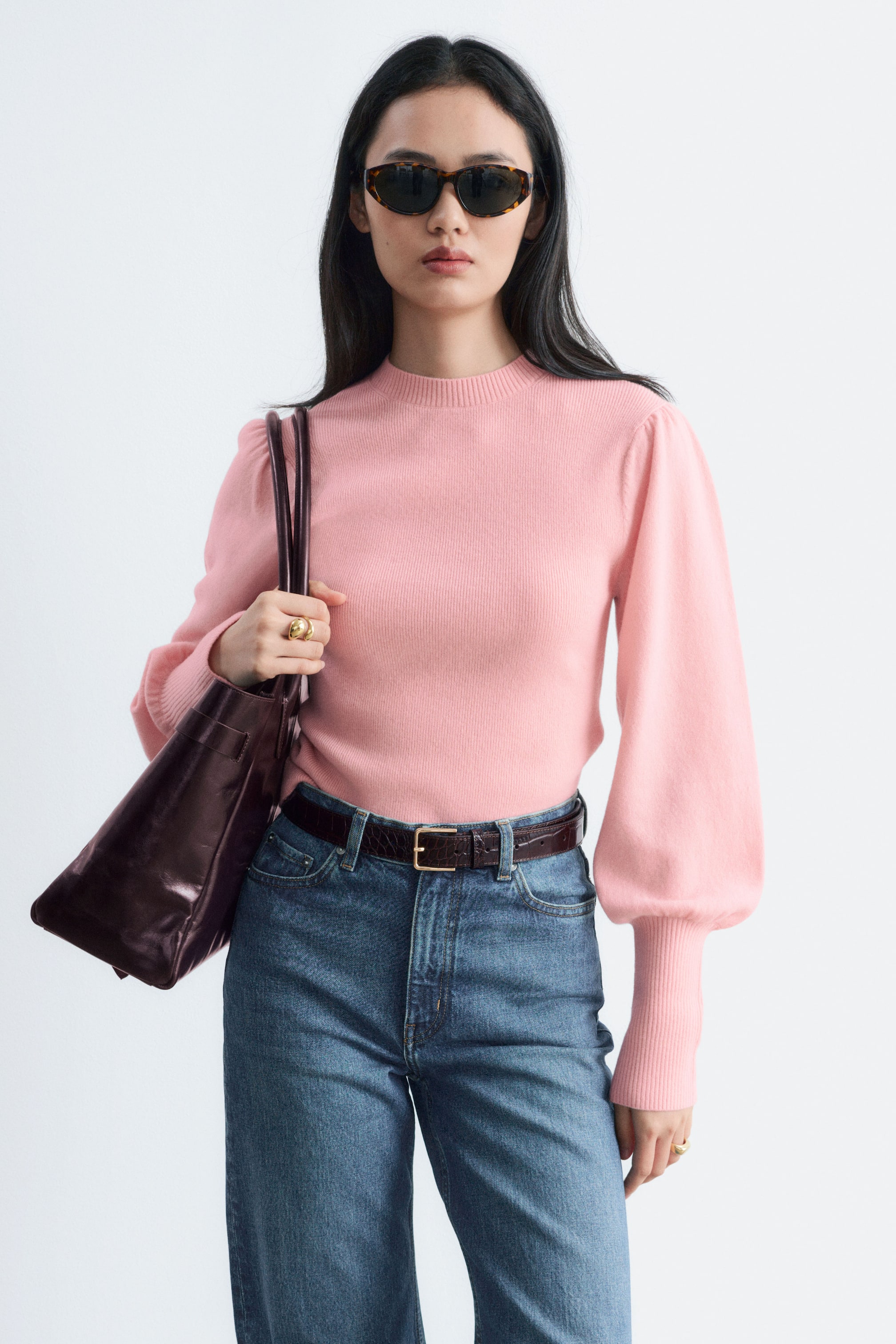 Grotere afbeelding bekijken: Het model draagt een lichtroze wollen top met een ribbel-mockneck en lange ballonmouwen met verlengde ribgebreide boorden. De top is ingestopt in een blauwe high-waist jeans, met een donkerleren totebag over één schouder gedragen.