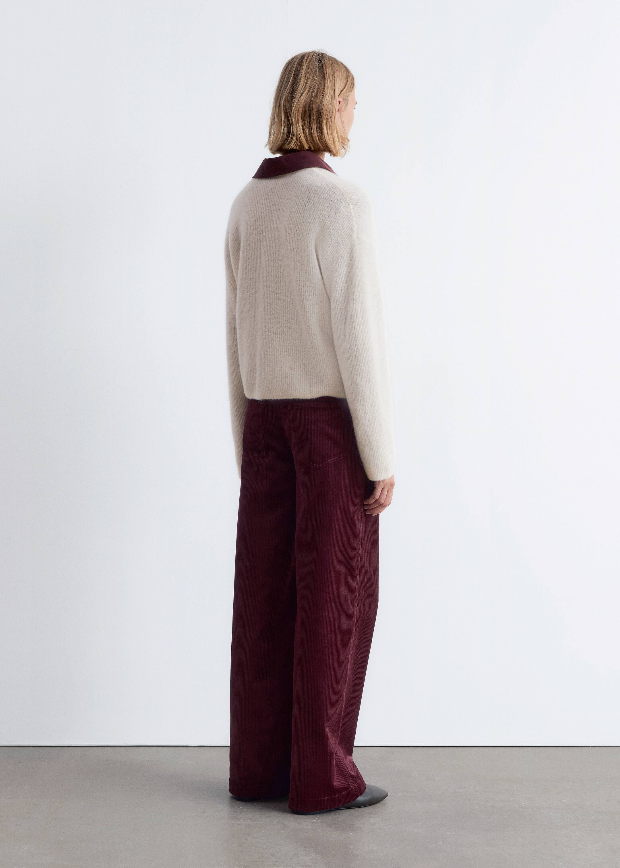 Visualizza immagine più grande: Un maglione color crema chiaro, a vestibilità rilassata, con maniche lunghe è indossato sopra pantaloni a gamba larga in velluto a coste rosso scuro, visto da un'angolazione posteriore di 3/4. Un colletto rosso scuro a contrasto è visibile sotto il maglione, e i pantaloni presentano una silhouette ampia con evidenti coste verticali in velluto a coste.