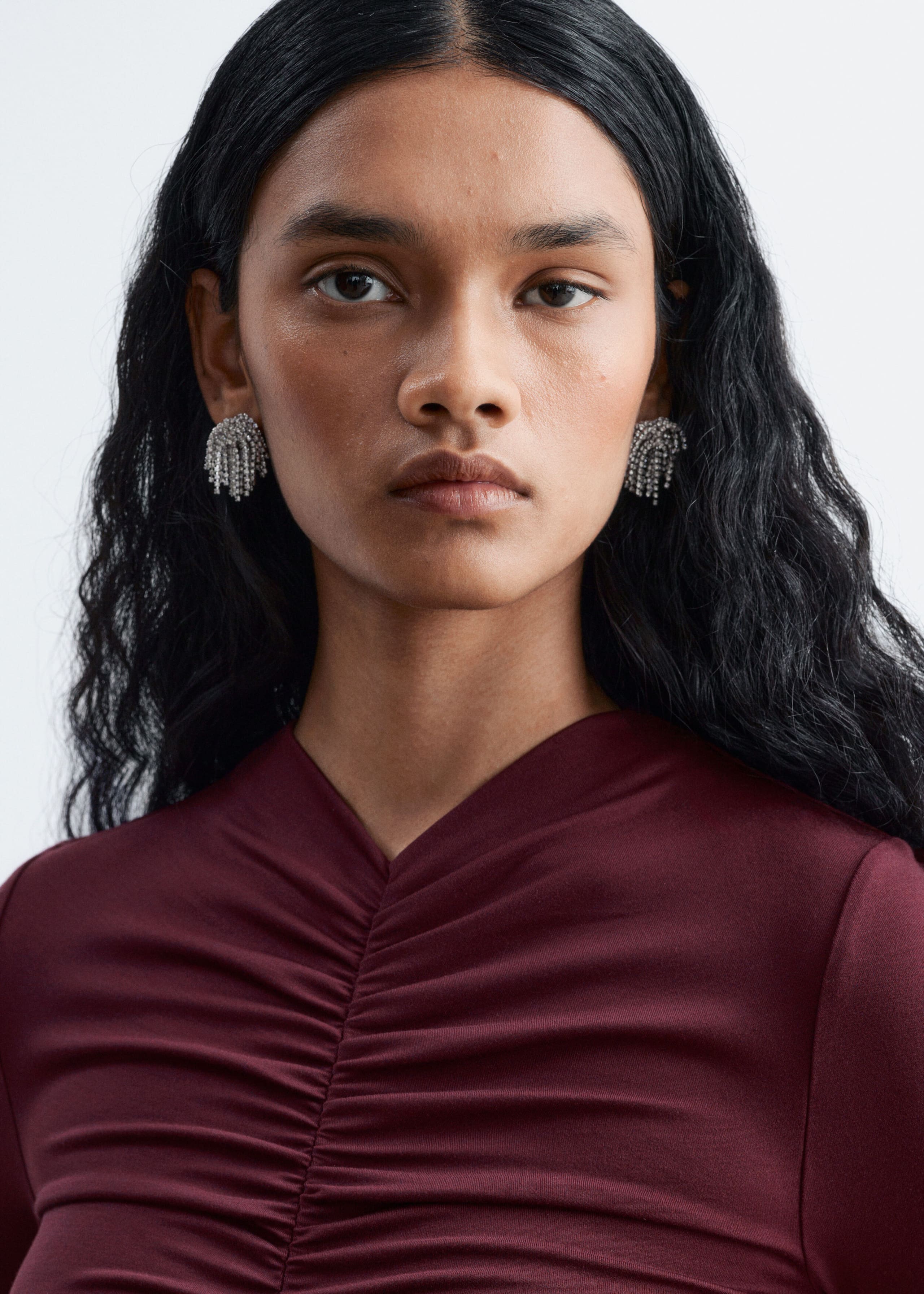 The model is wearing a burgundy jersey top featuring a gathered center-front seam. The ruching creates soft horizontal folds across the chest, shaping a subtle V-neckline.