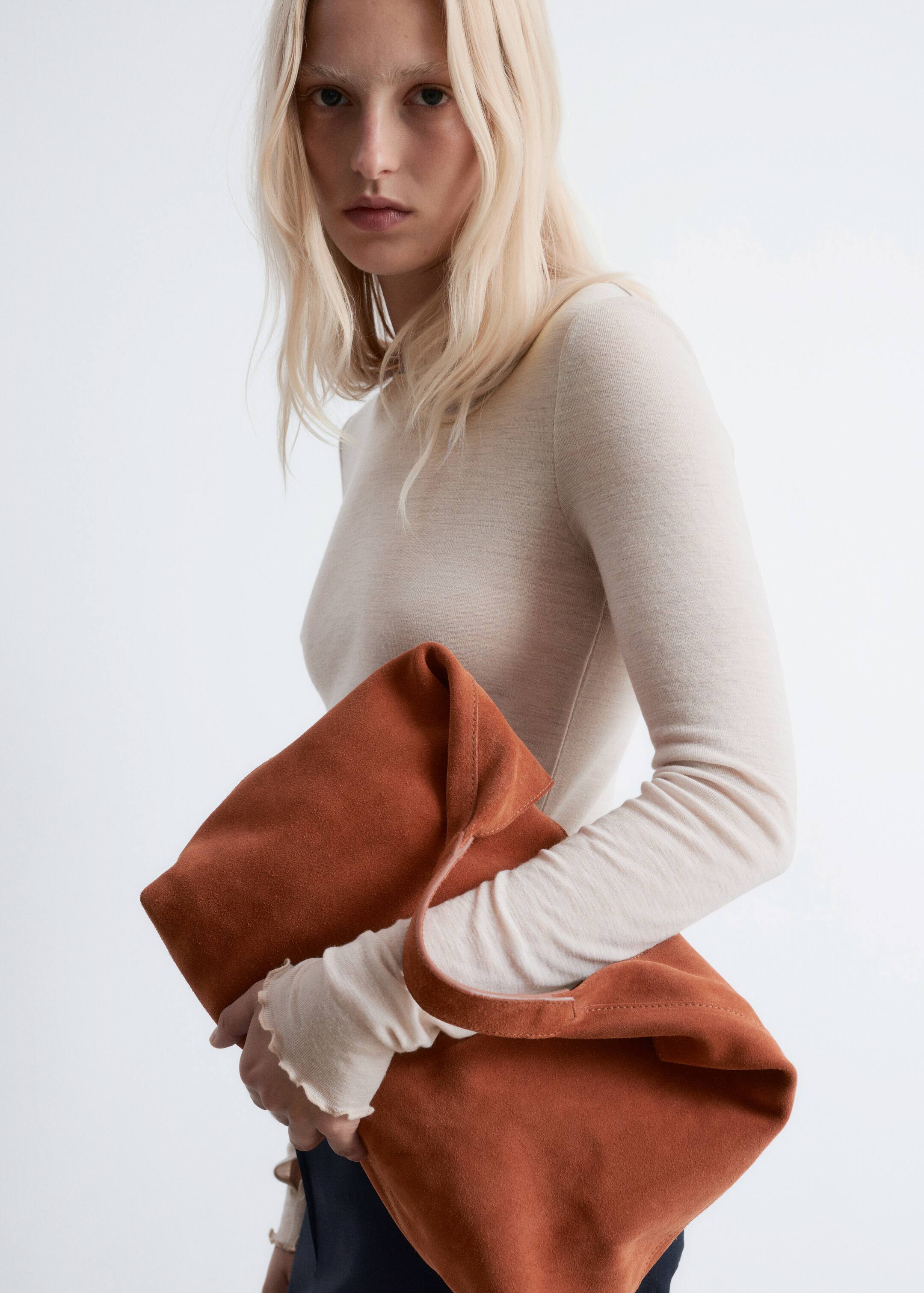Grösseres Bild ansehen: Eine weiche, unstrukturierte Tasche aus verbranntem orangefarbenem Wildleder wird schräg gehalten und zeigt ihr biegsames Material und den breiten, flachen Riemen, während sie sich natürlich faltet.