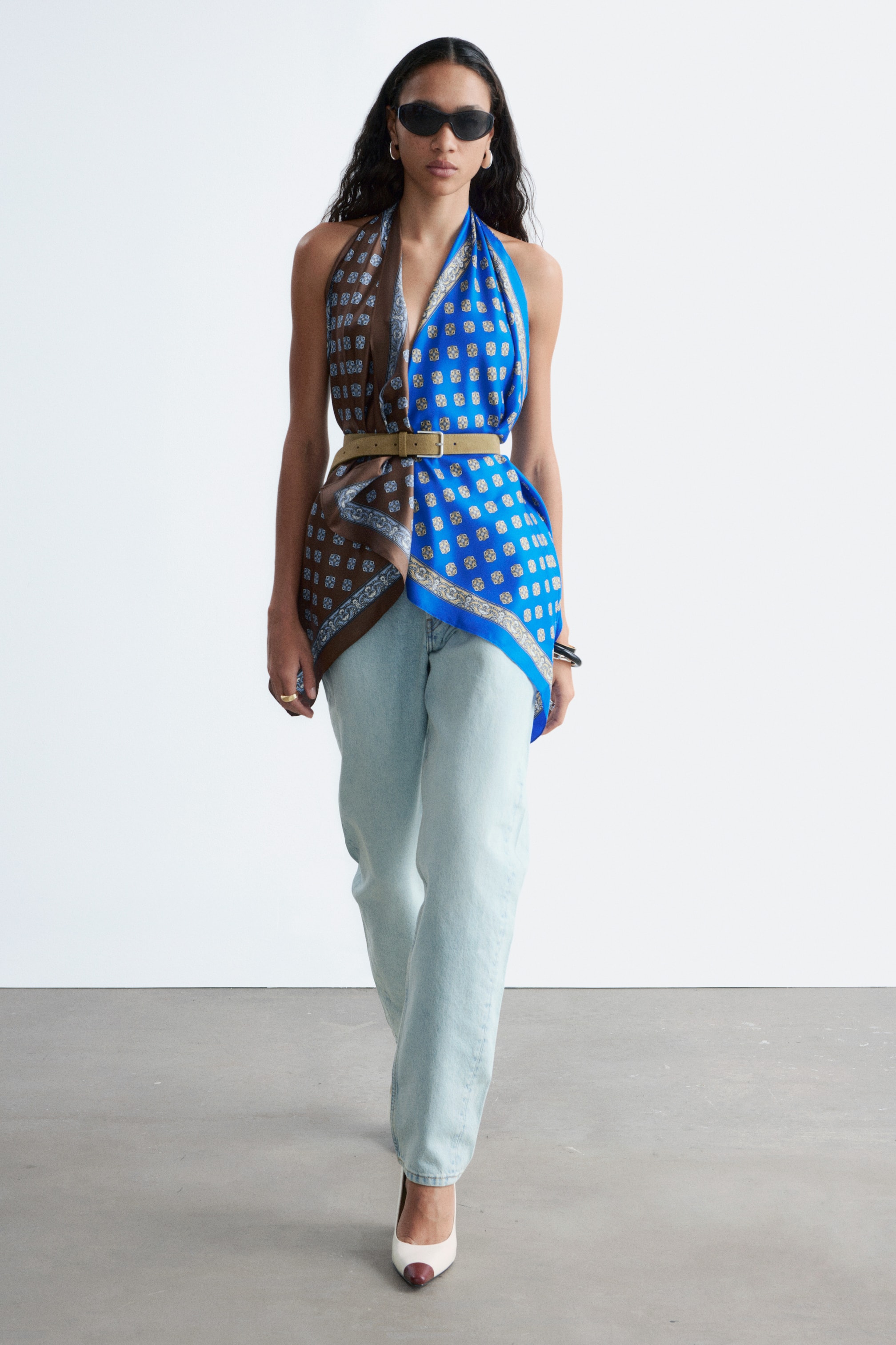Ver imagen más grande: Se presenta un top de pañuelo de frente, con cuello halter y escote en V con un dobladillo asimétrico. Muestra dos patrones distintos: uno con base marrón y otro con base azul, ambos con motivos geométricos. Un cinturón color tostado con una hebilla dorada rectangular ajusta el pañuelo en la cintura.