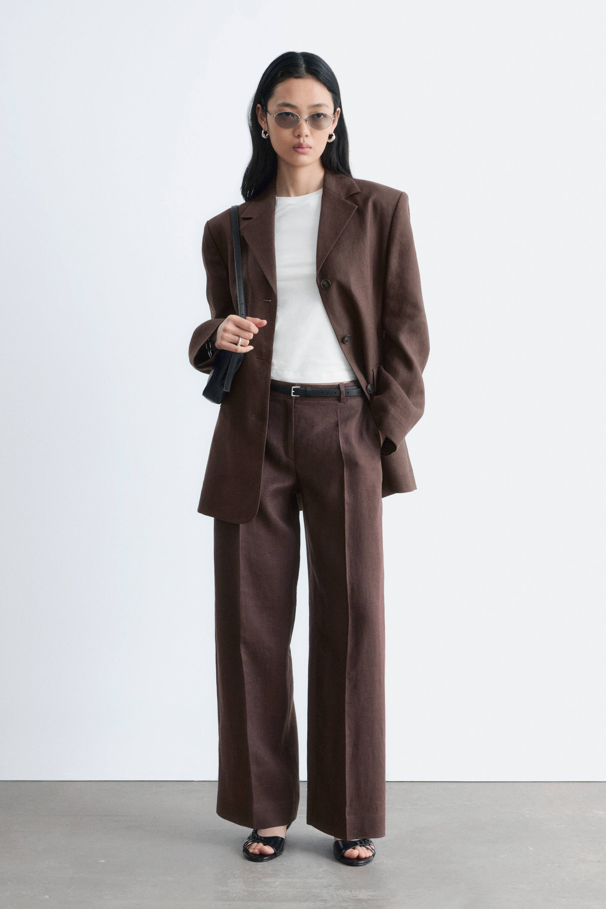 הצגת תמונה גדולה יותר: חליפה חומה עם כפתור אחד מוצגת, הכוללת בלייזר עם דשי חריץ, כתפיים מובנות וכיסי דש, שנלבשת פתוחה מעל חולצה לבנה. המכנסיים התואמים בעלי רגל רחבה מציגים חזית מקופלת ונקשרים במותן עם חגורה שחורה.