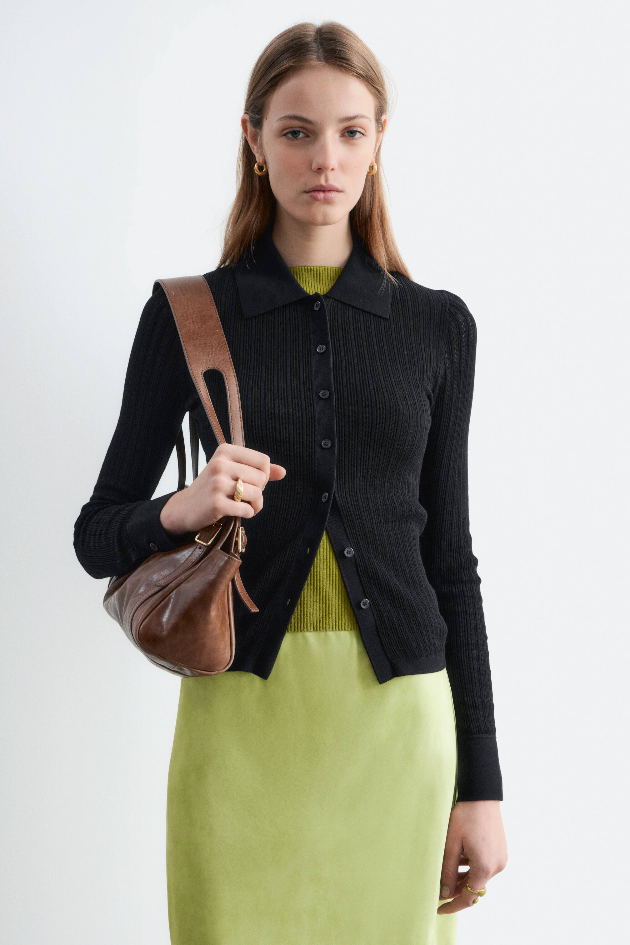 Größeres Bild ansehen: Die schwarze, gerippte Strickjacke zeichnet sich durch einen Polokragen und eine Knopfleiste mit dunklen Knöpfen aus. Sie ist mit langen Ärmeln gestaltet und wird offen über einem leuchtend grünen Top und Rock getragen, was ihr figurbetontes Design zur Geltung bringt.