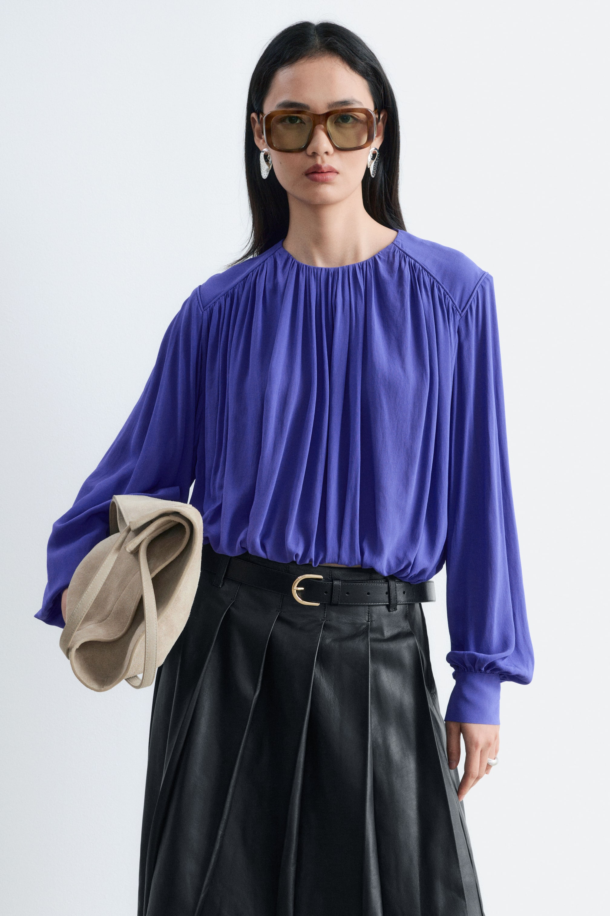 Visualizza immagine più grande: La modella indossa una blusa viola arricciata con scollo tondo e maniche lunghe con polsini. La blusa è infilata in una gonna nera plissettata con cintura in pelle, e una borsa beige è tenuta sotto il braccio della modella.