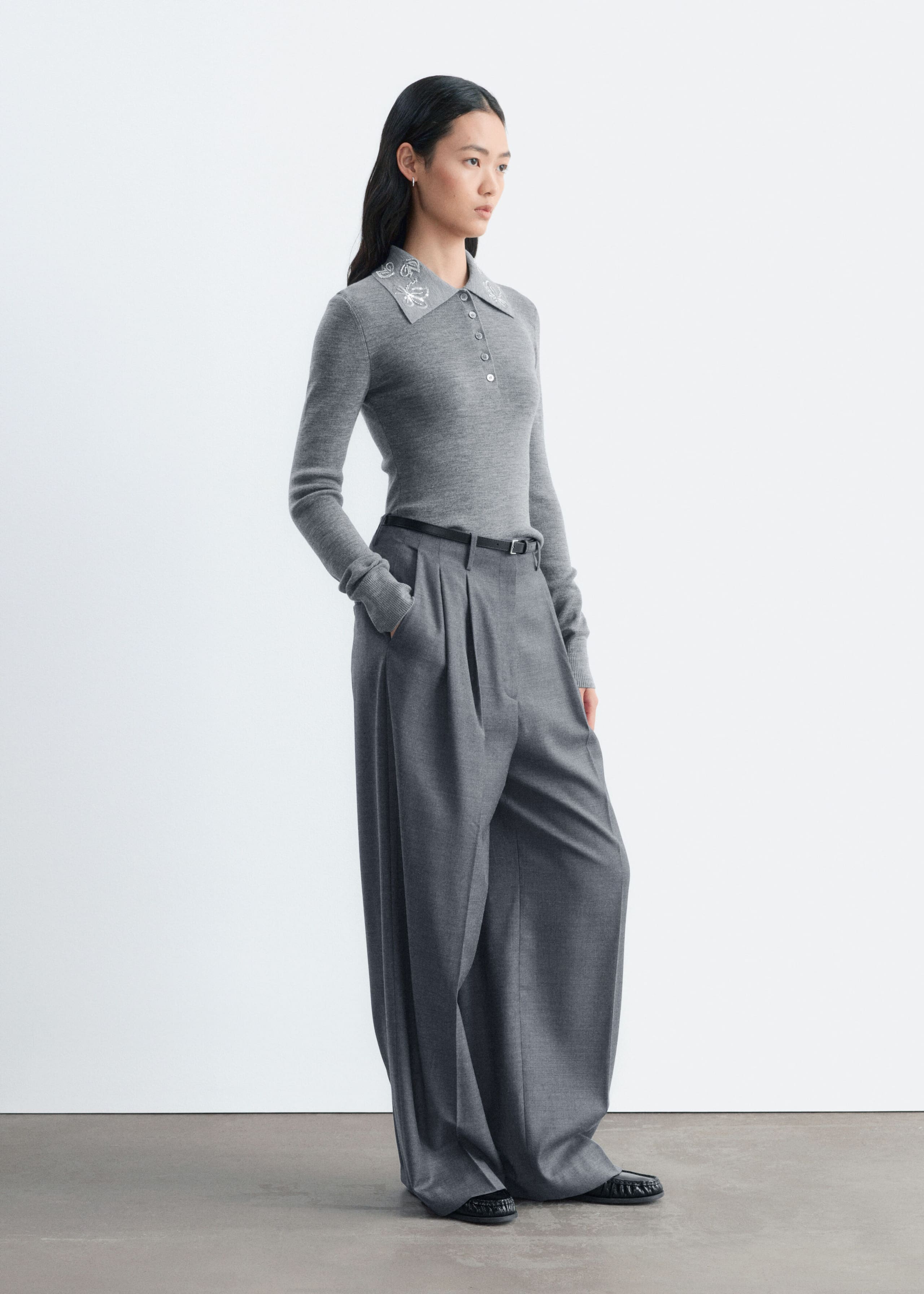 Ein grauer Woll-Pullover mit langen Ärmeln, Slim Fit, Knopfleiste und weisser Blumenstickerei am Kragen wird vom Model getragen. Der Pullover ist in die dazu passende graue Wide-Leg-Hose gesteckt, die mit einem schmalen schwarzen Gürtel gehalten wird.