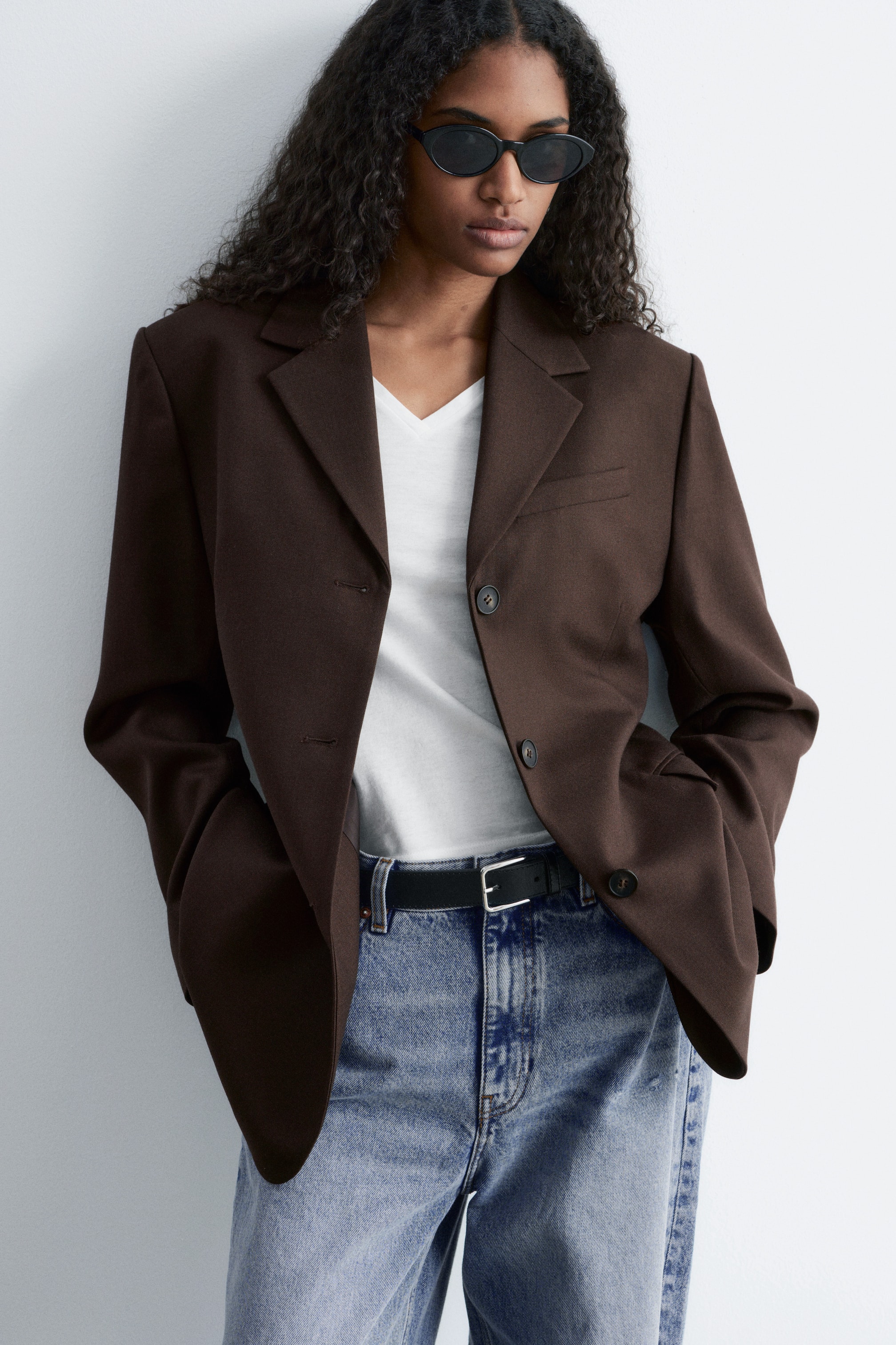 Vis større bilde: En brun blazer bæres av modellen, lagd over en hvit t-skjorte. Blazeren har strukturerte skuldre, hakkede slag, en brystlomme, to klafflommer og en to-knapps front som er åpen. Modellens hender hviler i klafflommene, og blazeren er stylet med blå jeans og et svart belte.