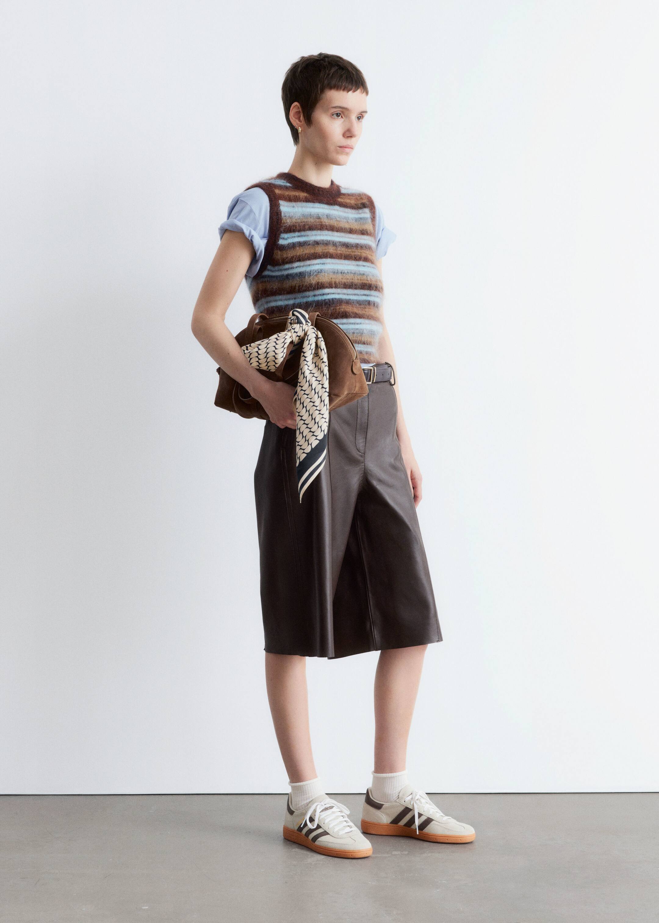 Größeres Bild ansehen: Eine Person trägt eine mehrfarbig gestreifte, flauschige Strickweste mit horizontalen braunen, hellblauen und cremefarbenen Streifen, die über einem hellblauen kurzärmeligen T-Shirt getragen wird. Sie tragen auch dunkelbraune Leder-Culottes und hellfarbene Sneakers mit dunklen Streifen, während sie eine braune Handtasche halten, die mit einem cremefarbenen gemusterten Schal drapiert ist.