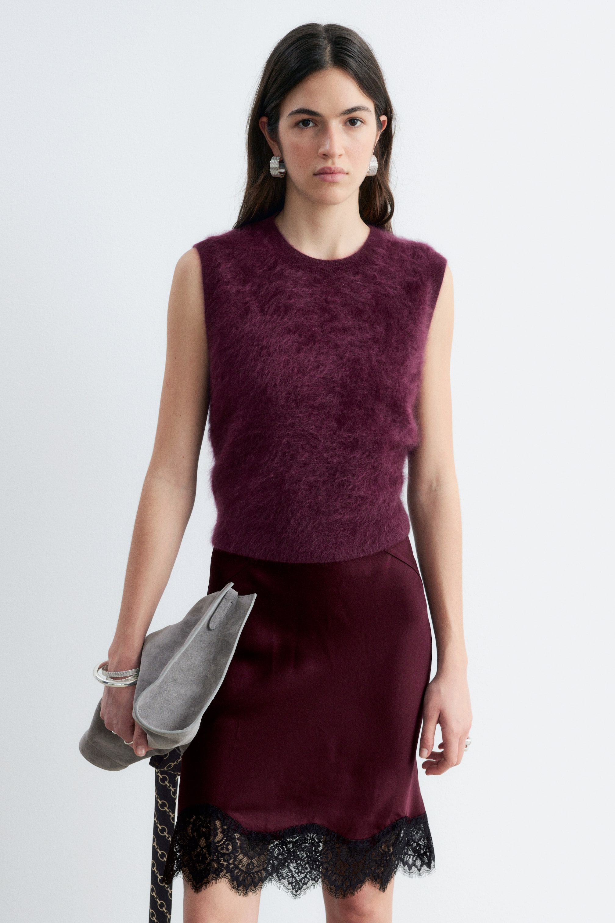 Visualizza immagine più grande: Un gilet in maglia senza maniche, color prugna intenso, in cashmere spazzolato con texture morbida e scollo tondo, indossato dalla modella. Il gilet è abbinato a una gonna in raso bordeaux coordinata, impreziosita da un bordo in pizzo nero.
